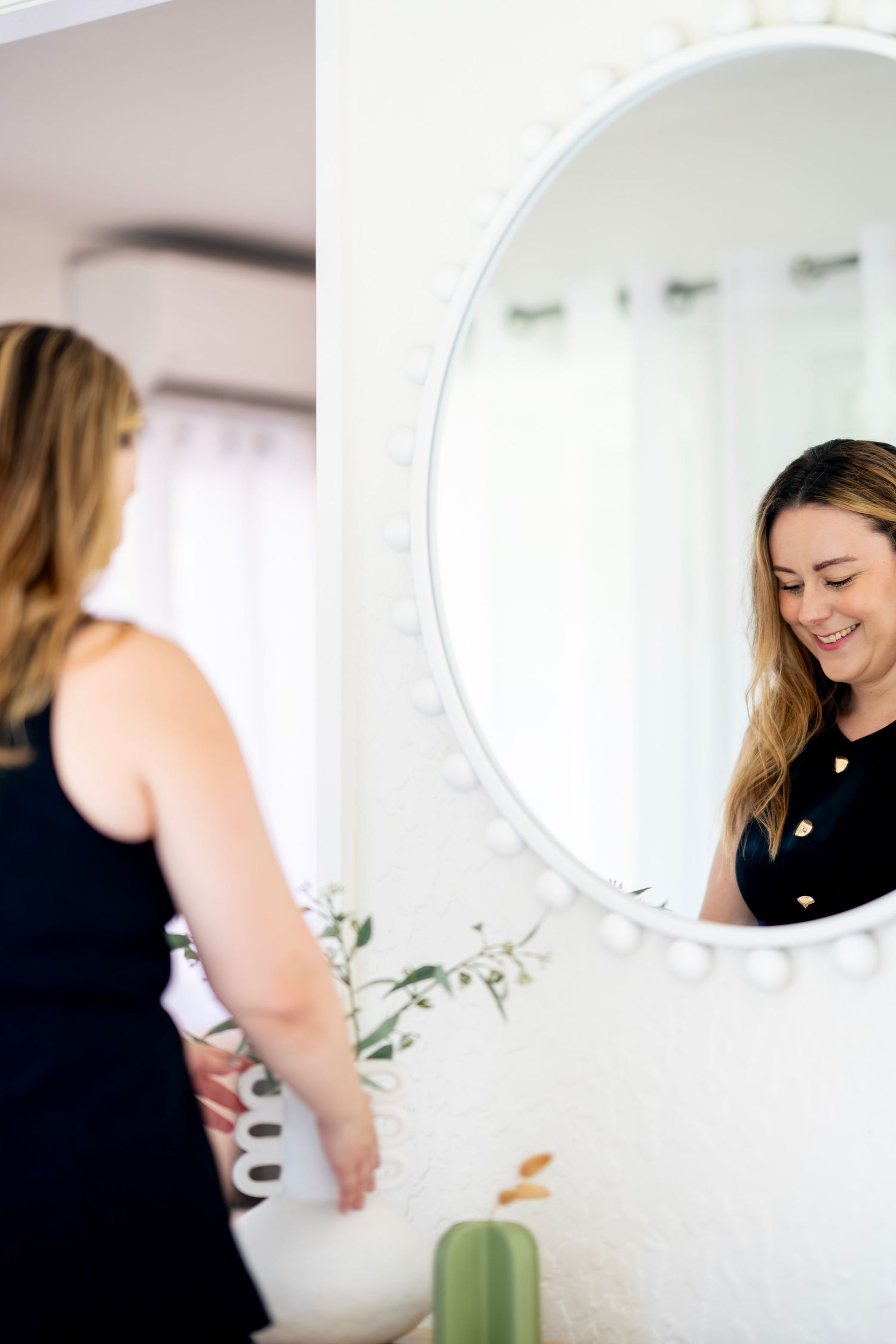 A smiling young woman in a black top looks at her reflection in a round white decorative mirror.