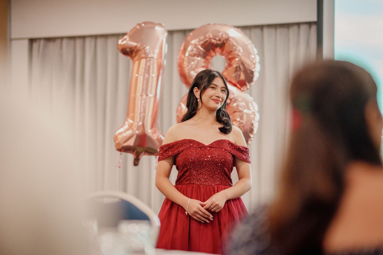 Young woman in red off-shoulder gown smiles at her 18th birthday party with rose gold number balloons behind her.