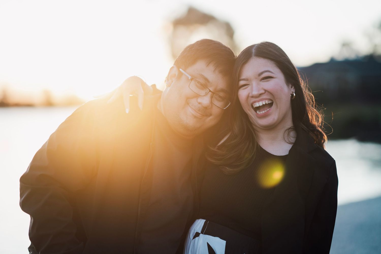 A joyful couple laughs together bathed in golden sunset light near a waterfront.