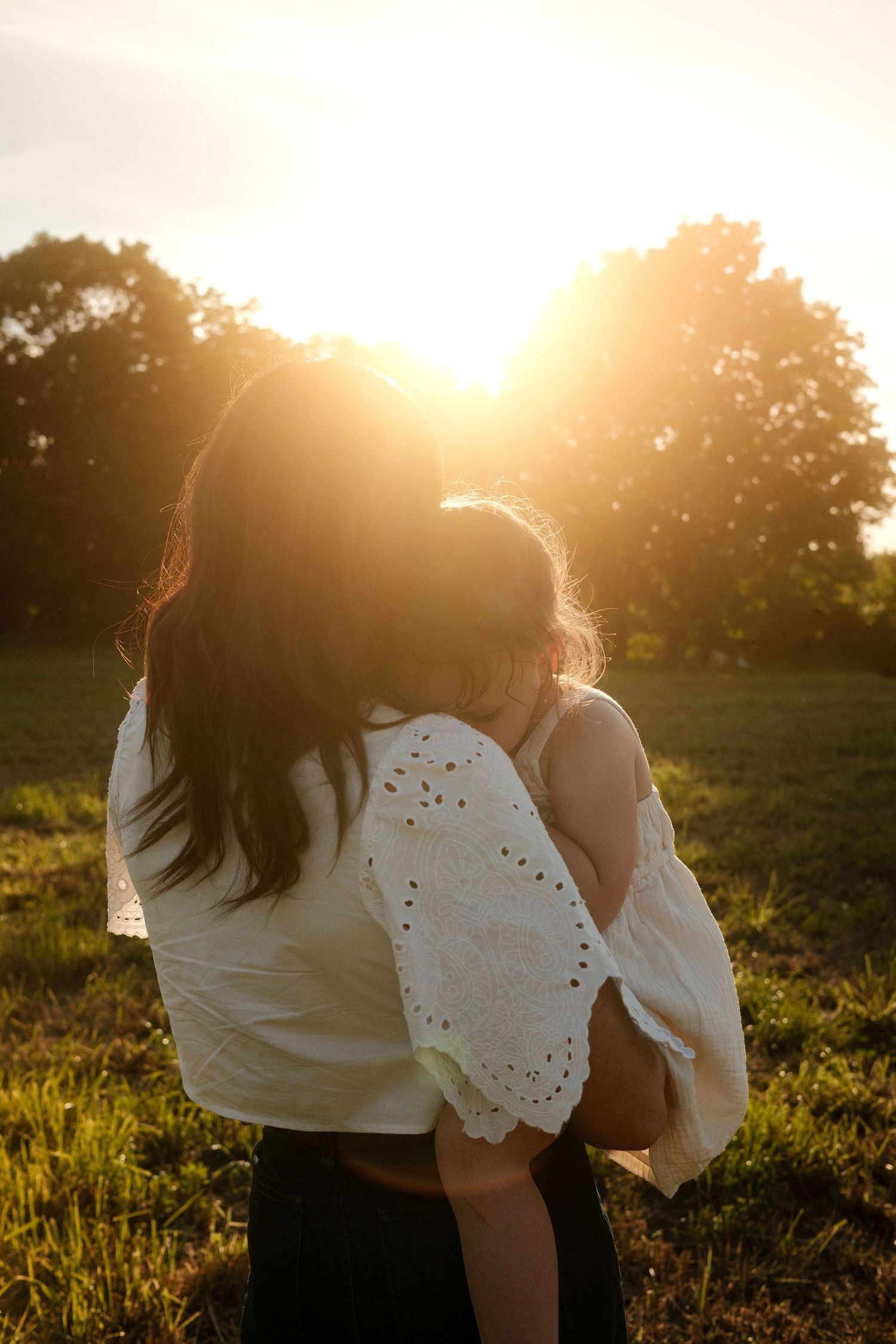 Mother holding child backlit by golden sunset in green field with trees.