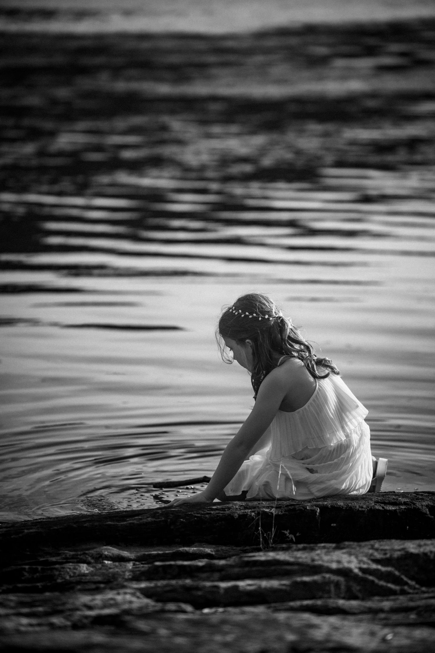 Black and white photo of a girl in white dress sitting by water's edge, touching the surface of a calm lake.