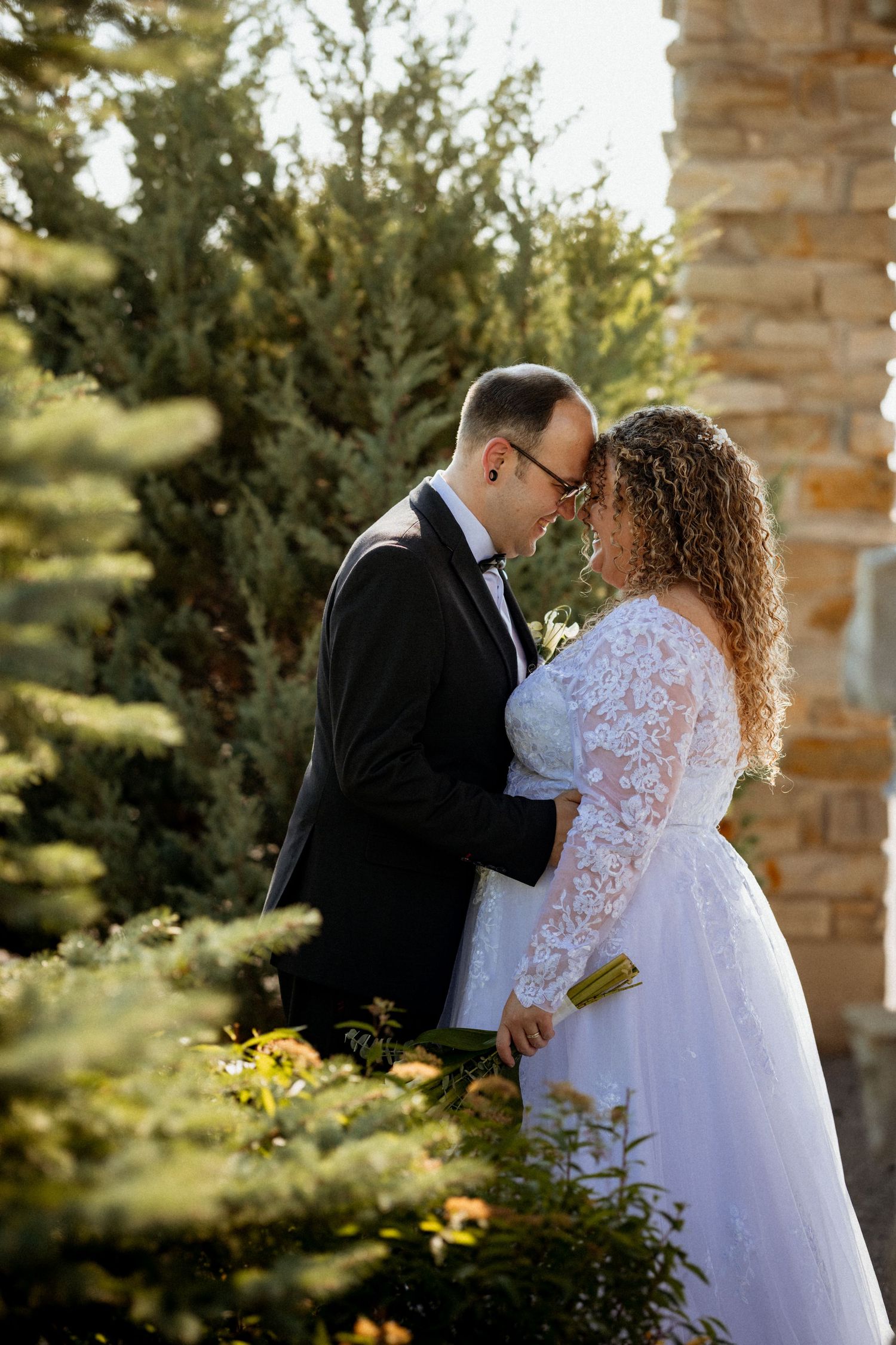 A bride and groom share a romantic forehead touch in a lush garden setting, with the bride in lace gown and groom in tuxedo.