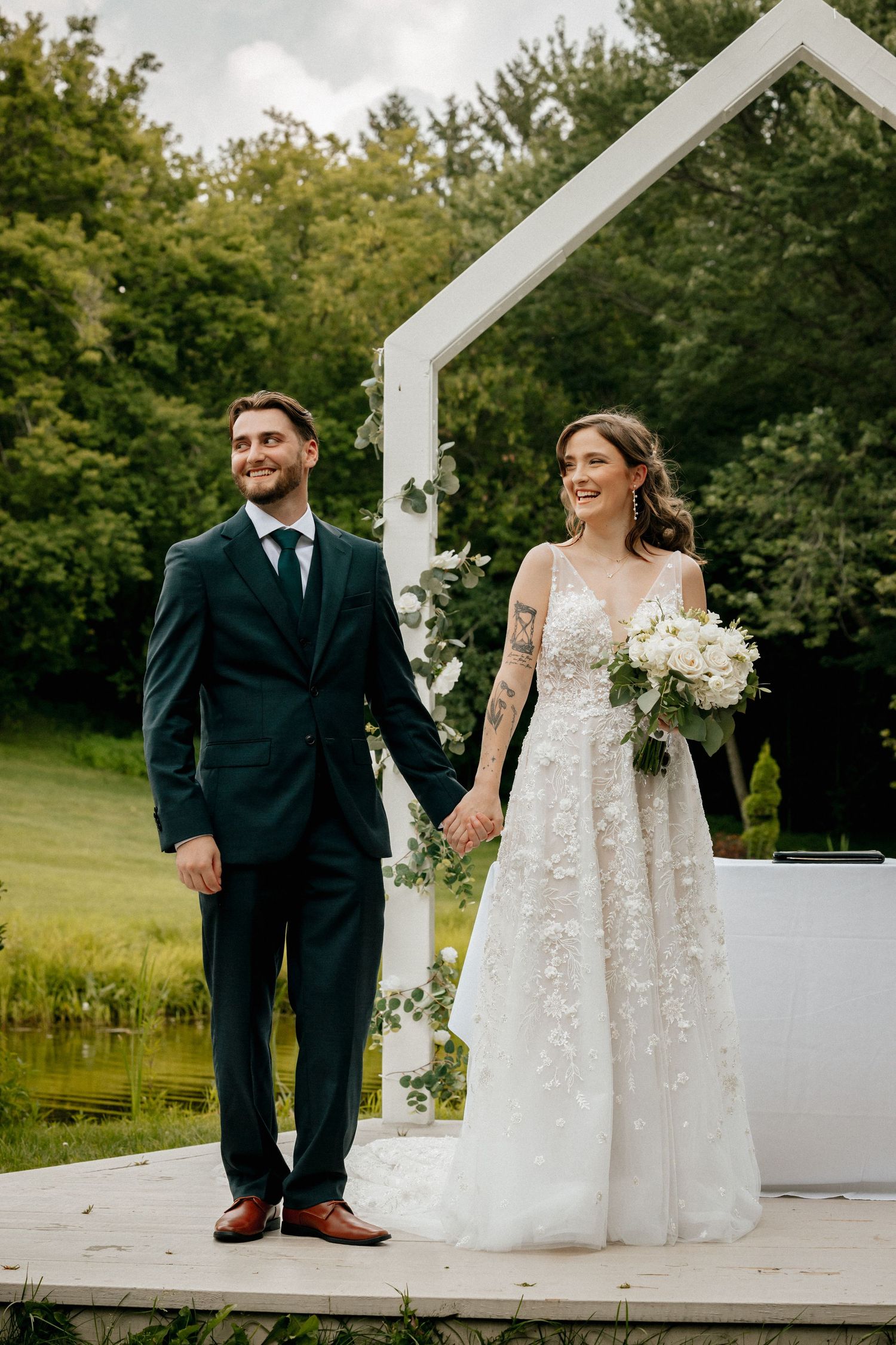 A smiling bride and groom hold hands outdoors near a white arch, surrounded by lush greenery on their wedding day.