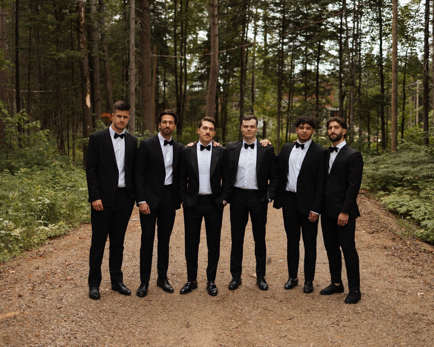 Six groomsmen in black tuxedos with bow ties posing on a forest dirt path surrounded by tall pine trees.