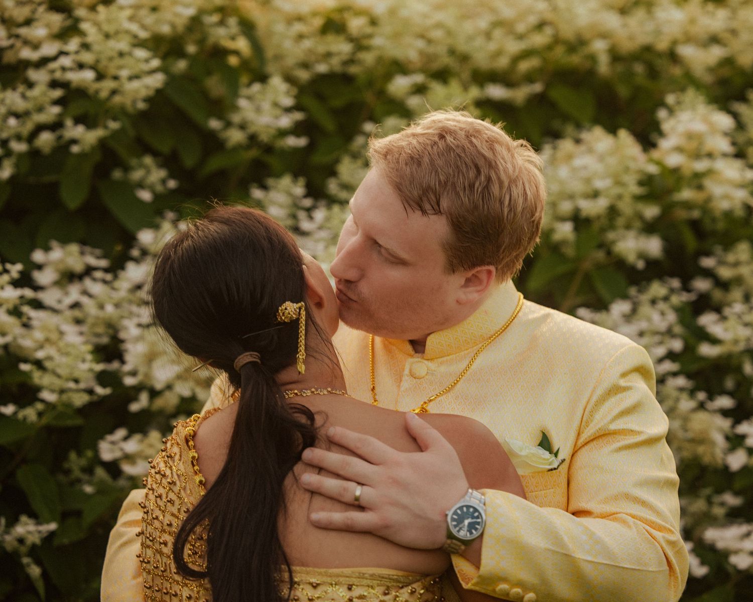 Couple shares a kiss surrounded by white flowers, groom in yellow traditional suit embracing bride in matching outfit.