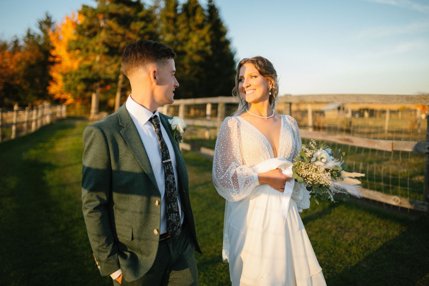 Bride holding bouquet smiling at groom during golden hour outdoor farm wedding portrait.