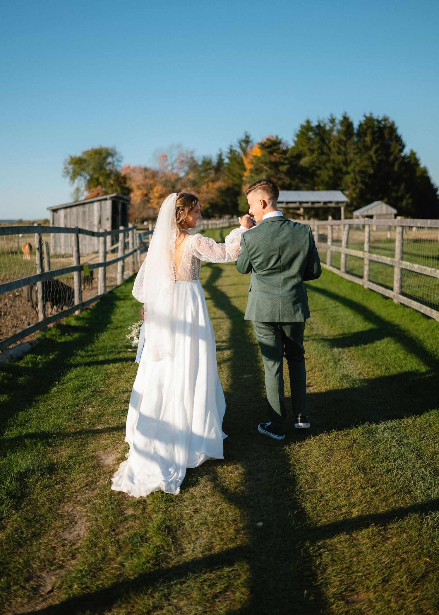 Bride and groom walking hand in hand along farm fence on sunny wedding day.