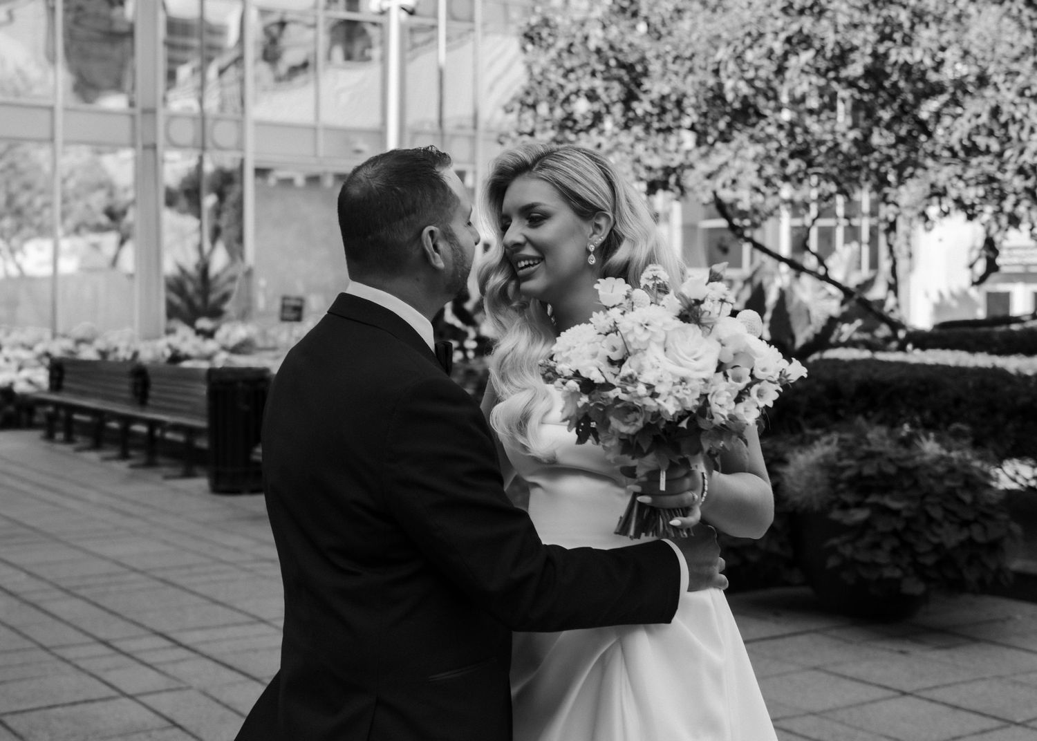 Black and white photo of a bride holding flowers smiling at her groom in a suit outdoors.