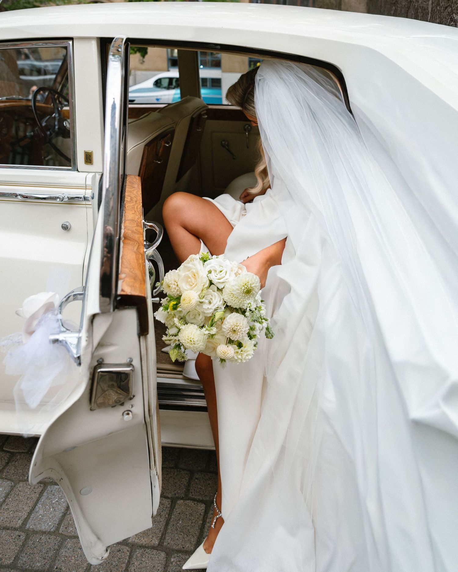 Bride in white gown and veil holding white bouquet stepping into vintage classic wedding car.