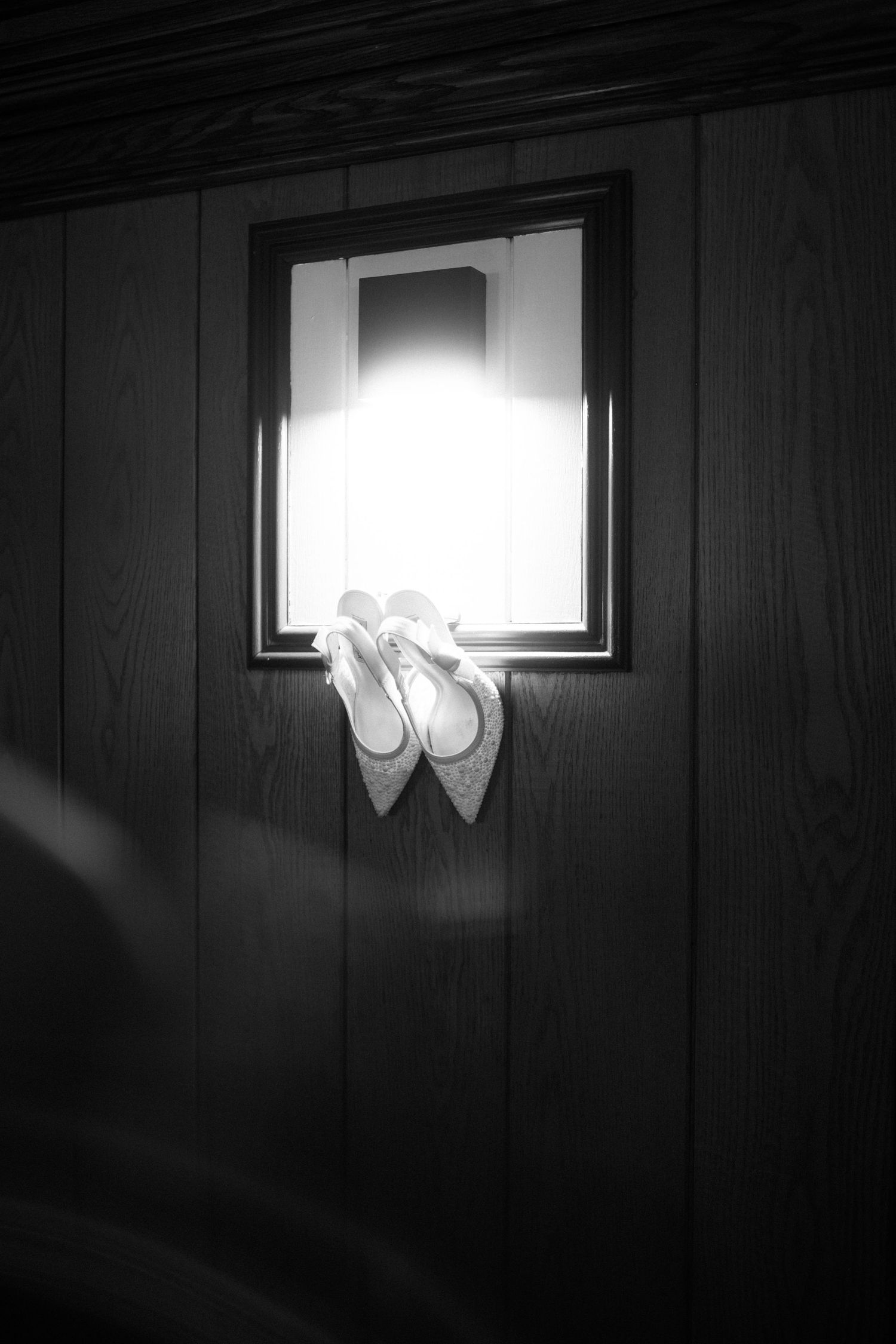Black and white photo of bridal heels displayed on a glowing window sill in a dark wood-paneled room.