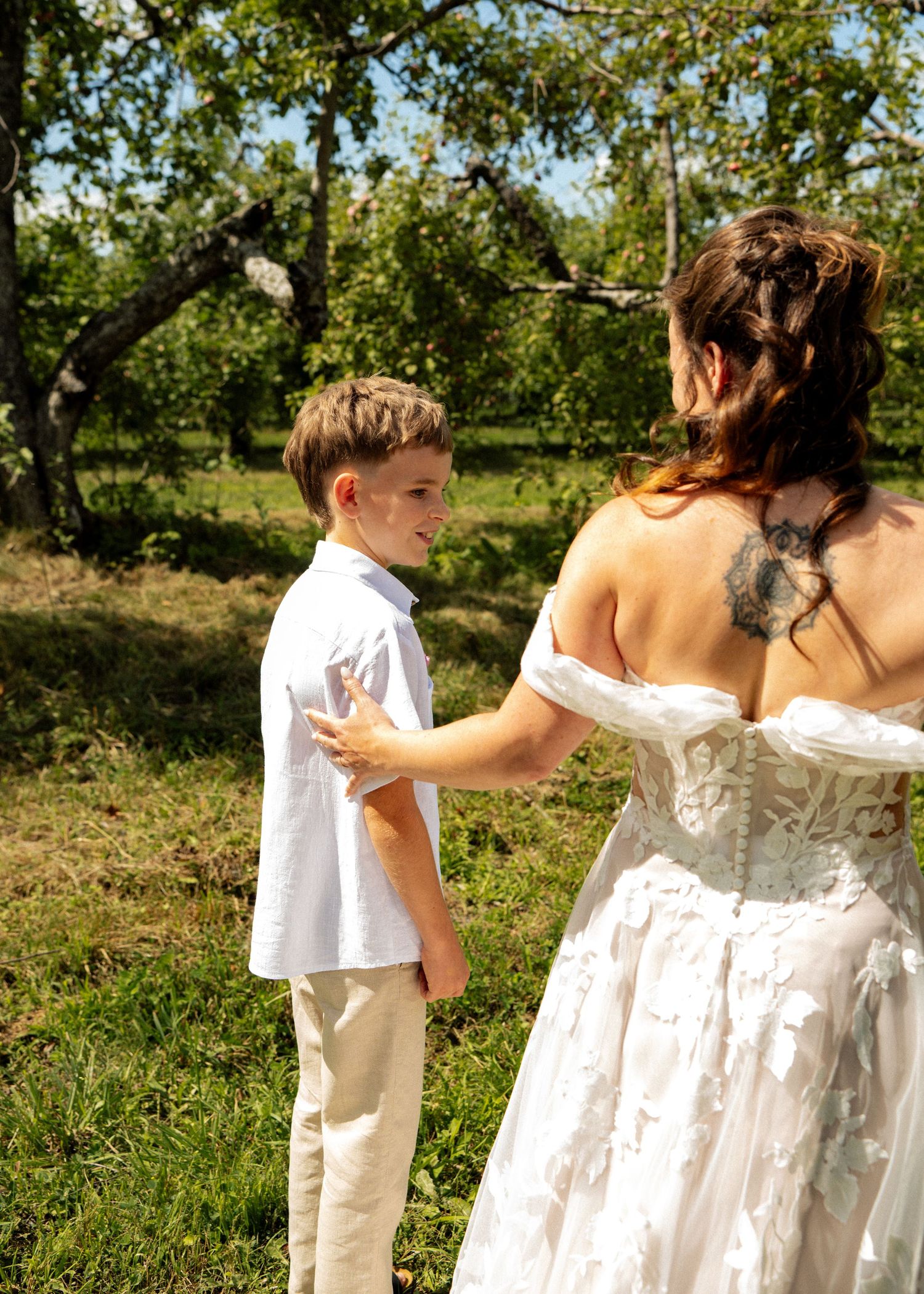 Young boy in white shirt holds hands with a bride in a lace wedding dress in a sunny orchard.
