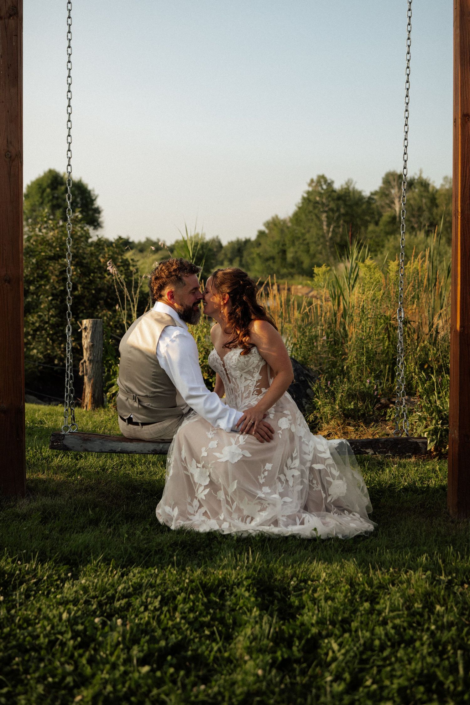 A bride and groom sit closely together on a wooden swing at golden hour, sharing an intimate moment in a lush garden setting.