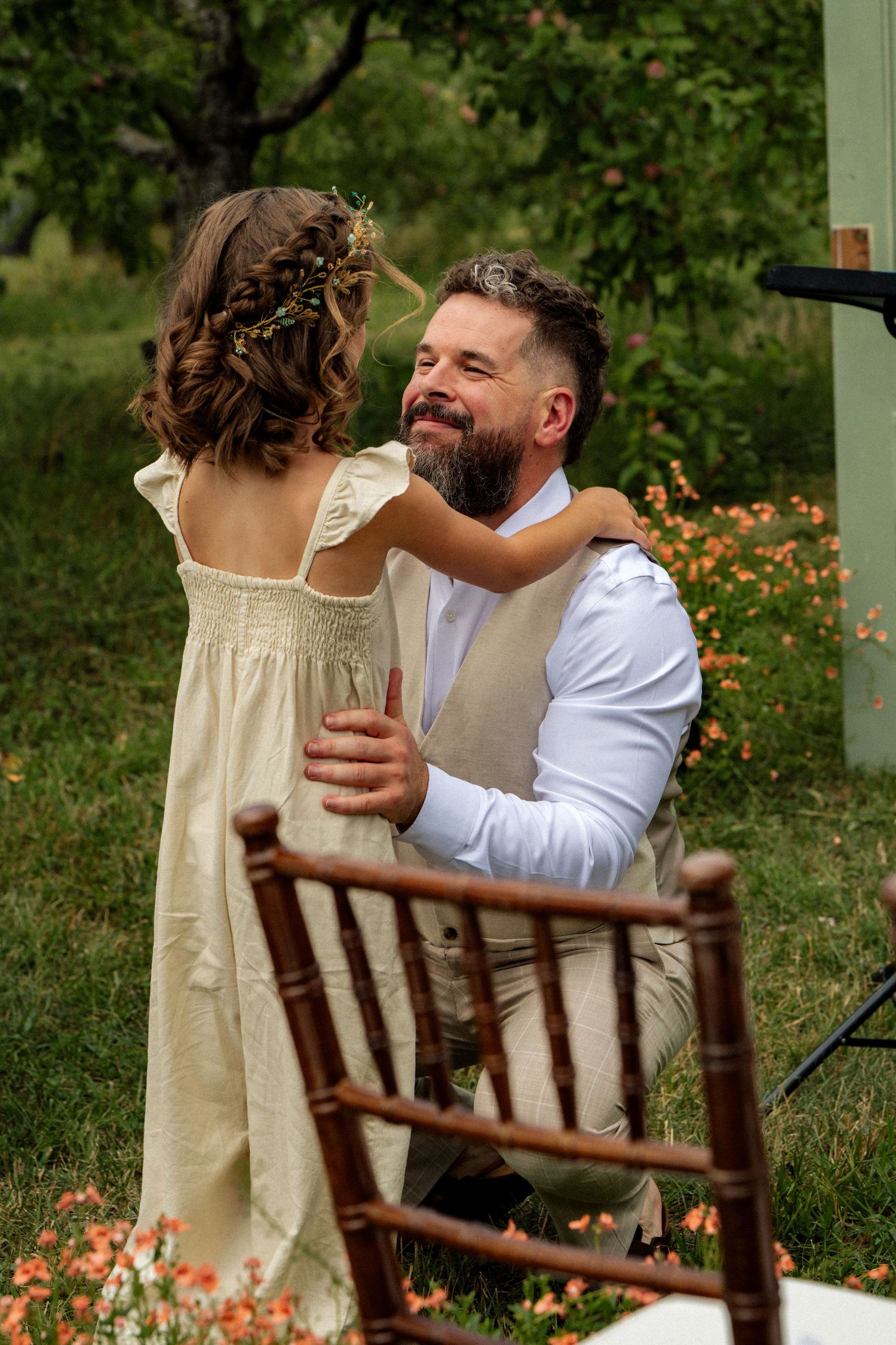 Bearded groom kneeling to embrace a flower girl outdoors.