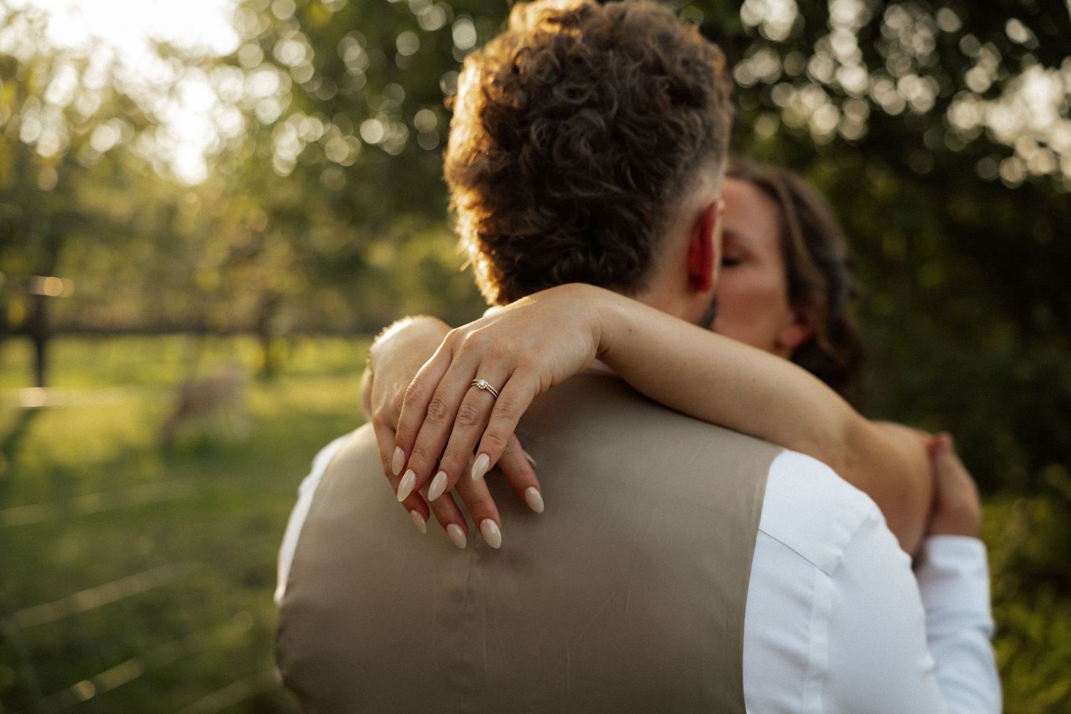 Bride with gold wedding ring embracing groom in sunlit green garden setting from behind.