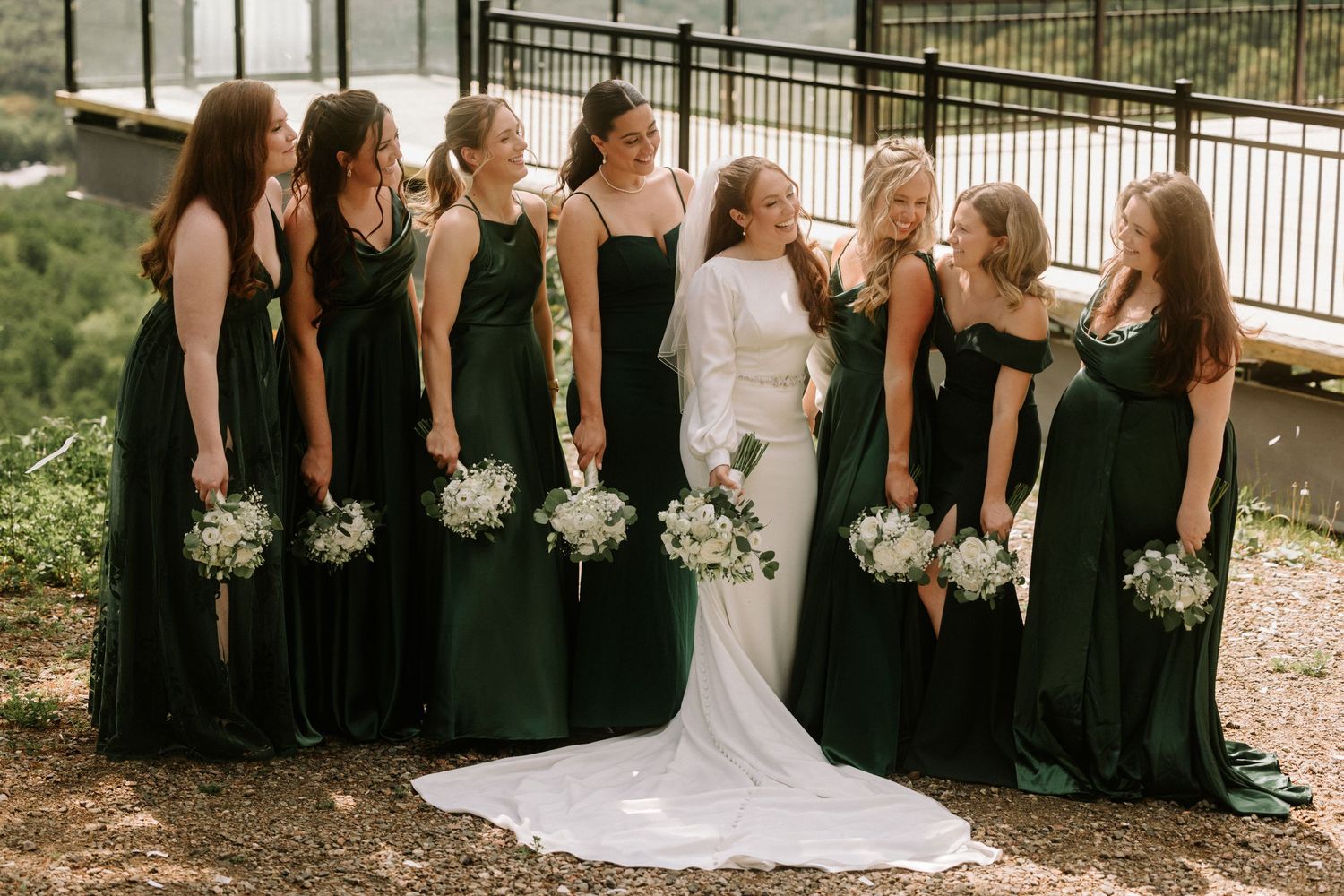 Bride in white gown with bridesmaids in emerald green dresses holding white bouquets outdoors.
