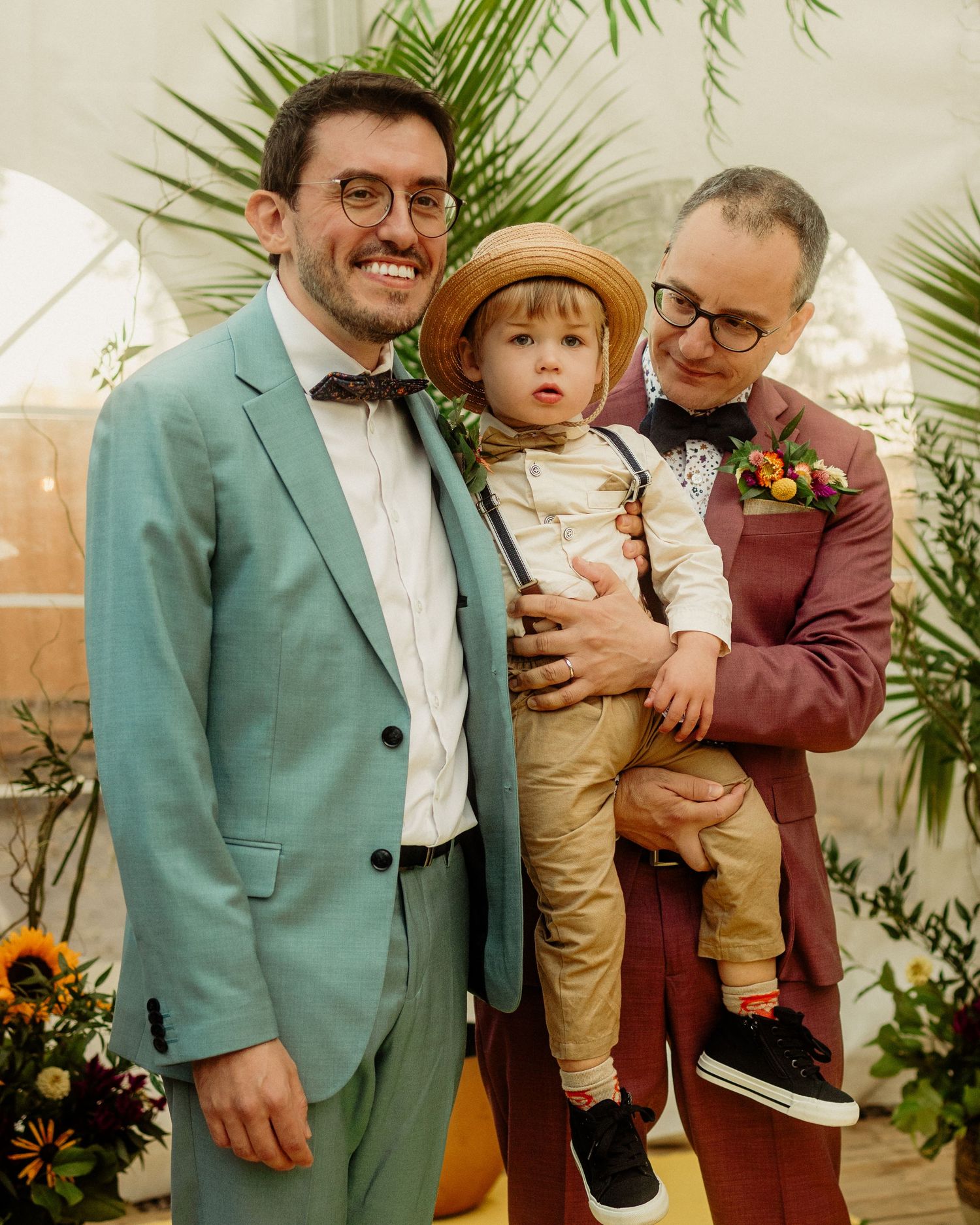 Two grooms in colorful suits pose with their young child in a straw hat at their wedding.