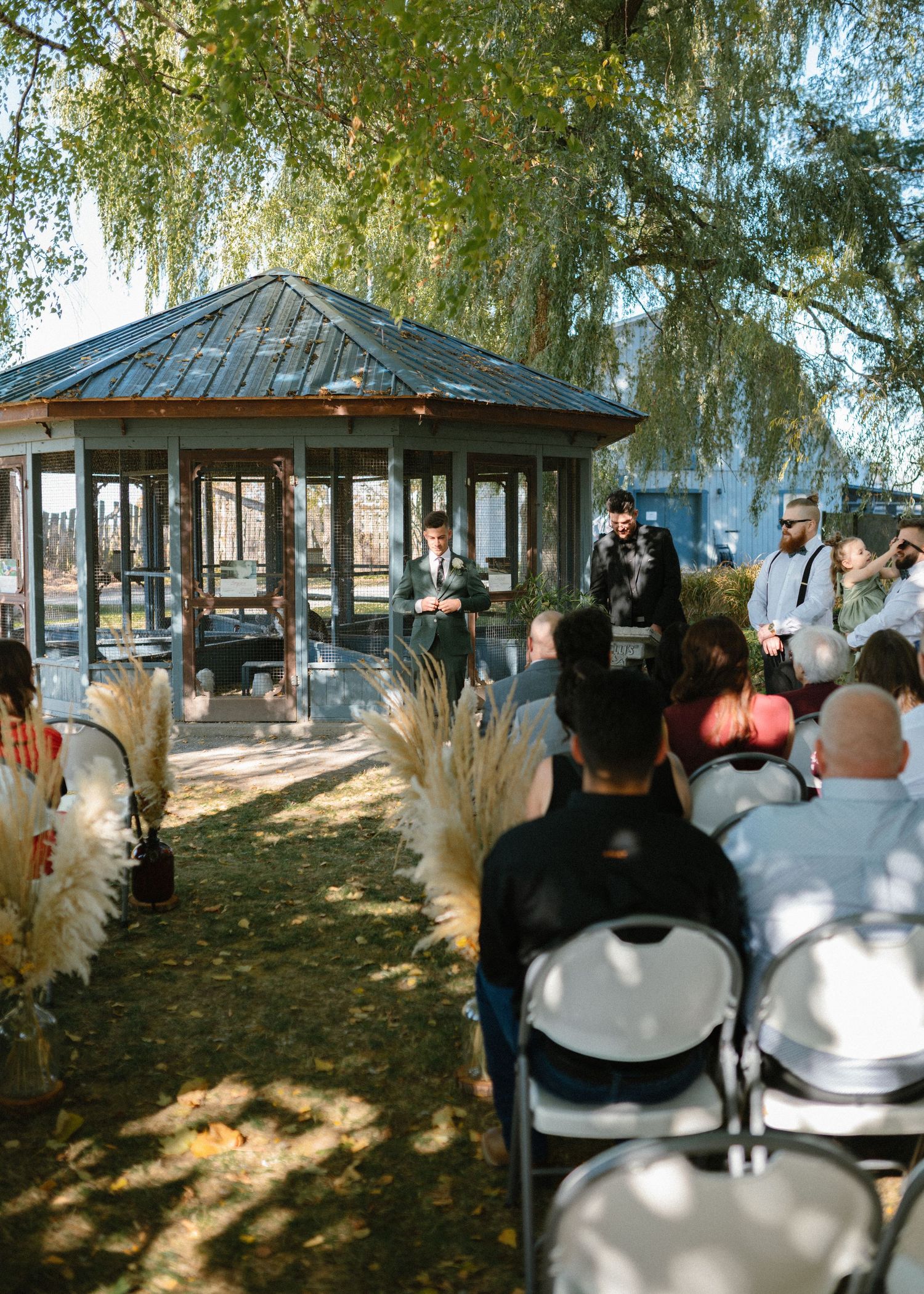Outdoor wedding ceremony with guests seated in white chairs facing a gazebo surrounded by willow trees.