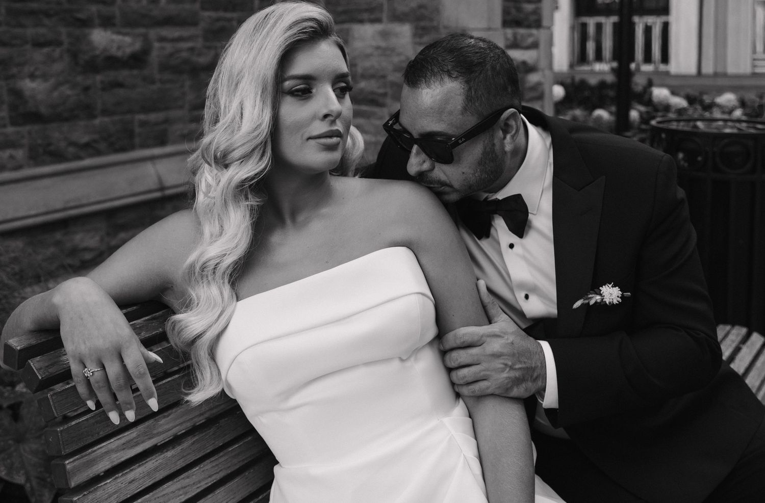 Bride in strapless white gown and groom in black tuxedo share an intimate moment on a bench.