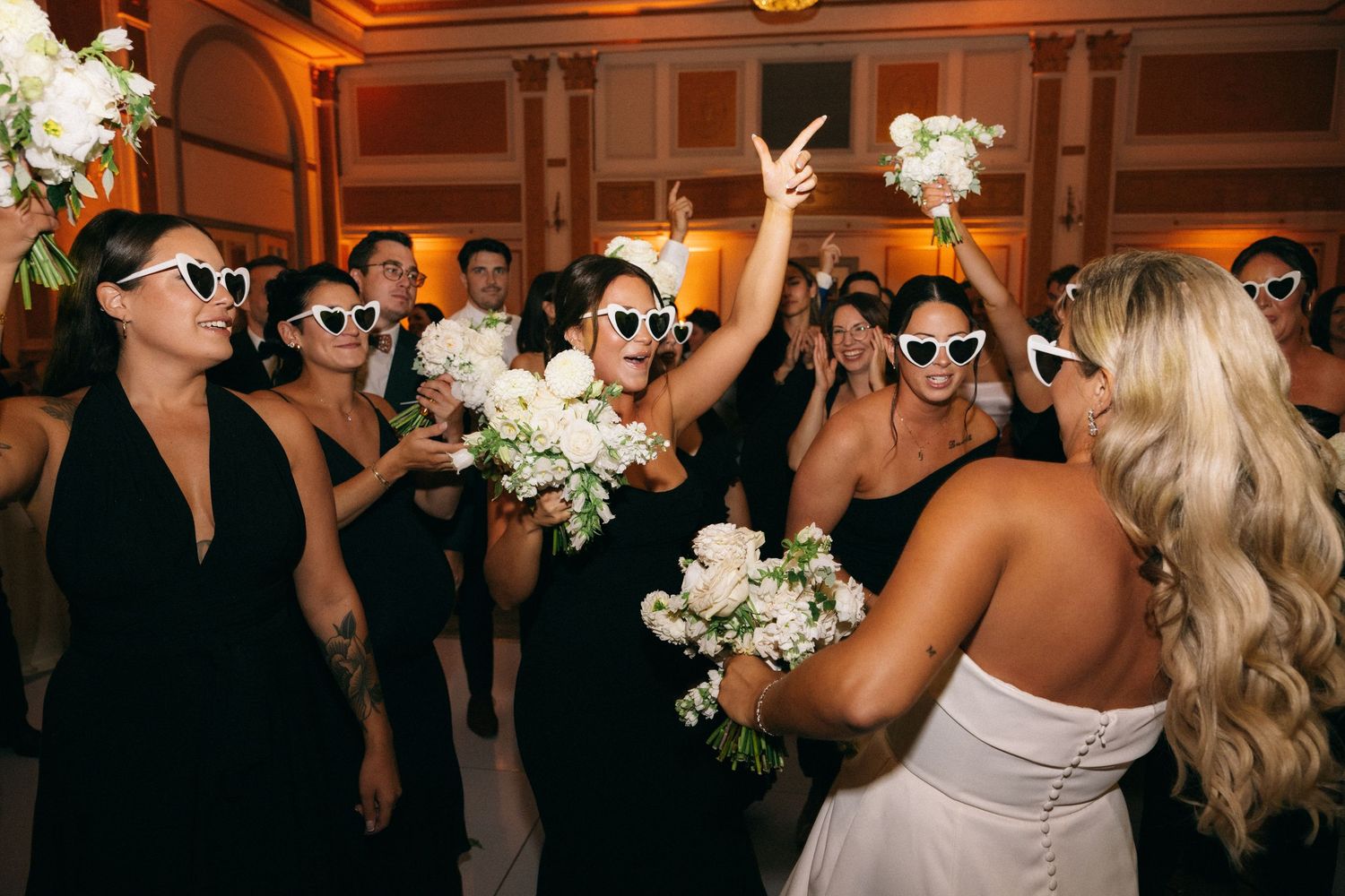Bridesmaids in black dresses and white heart sunglasses celebrating bouquet toss at reception.