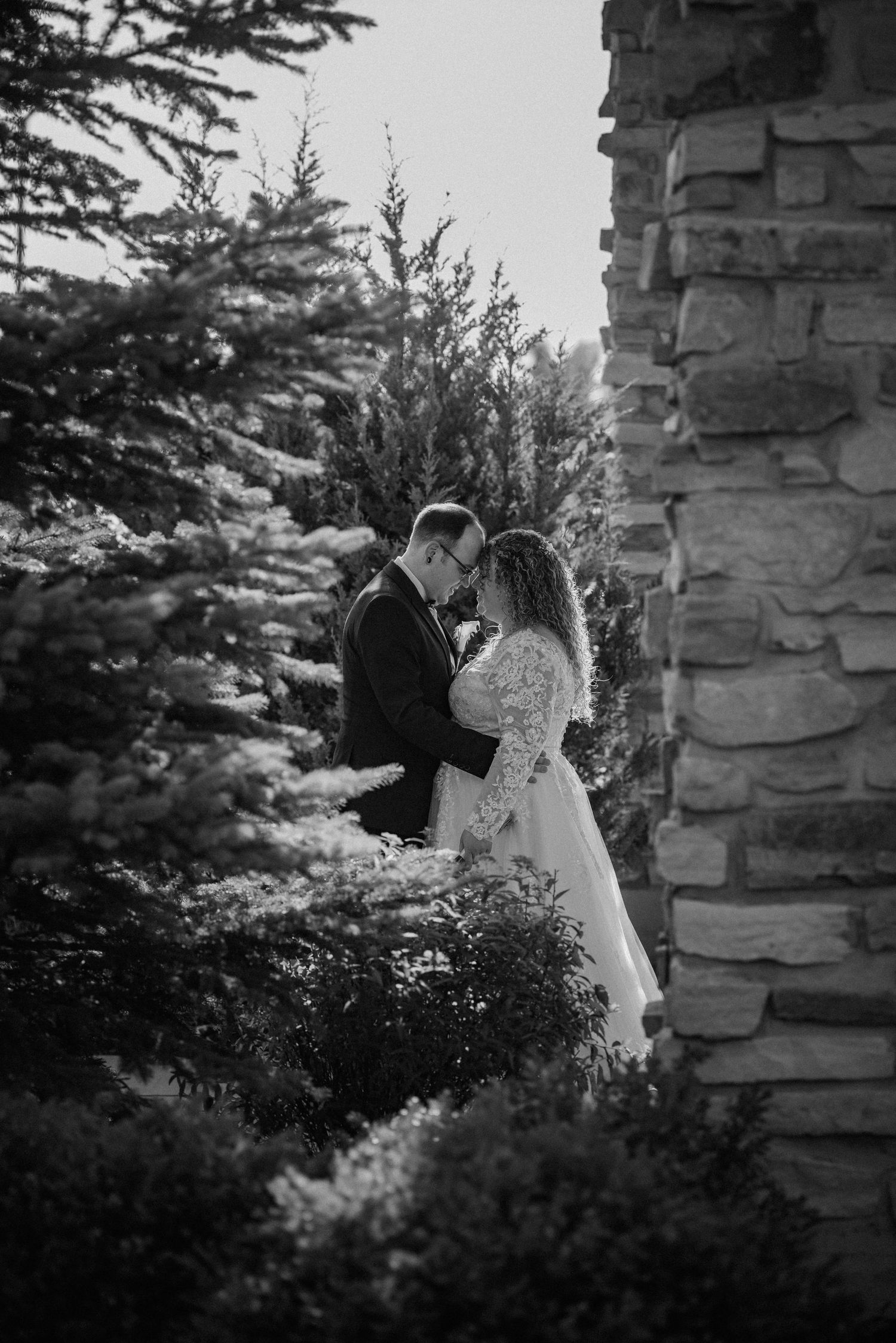 Black and white photo of bride and groom embracing near evergreen trees and rustic stone wall.