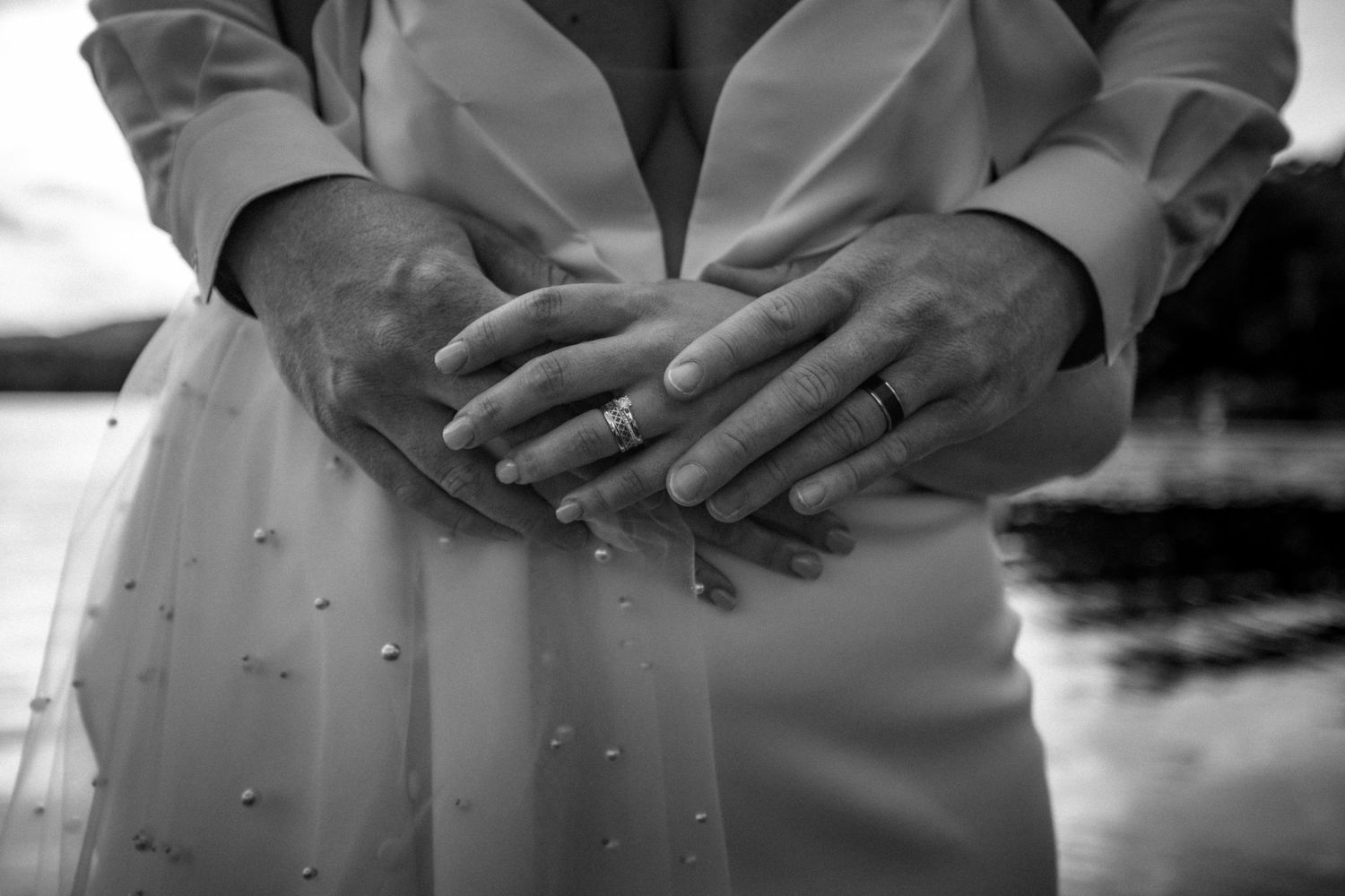 Black and white photo of newlyweds hands showing wedding rings on brides white pearl-adorned dress.