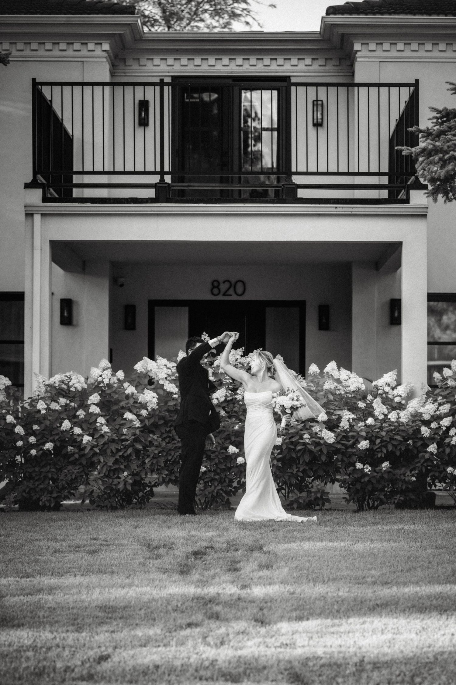 Black and white photo of bride and groom dancing in front of elegant building 820, surrounded by blooming flowers.