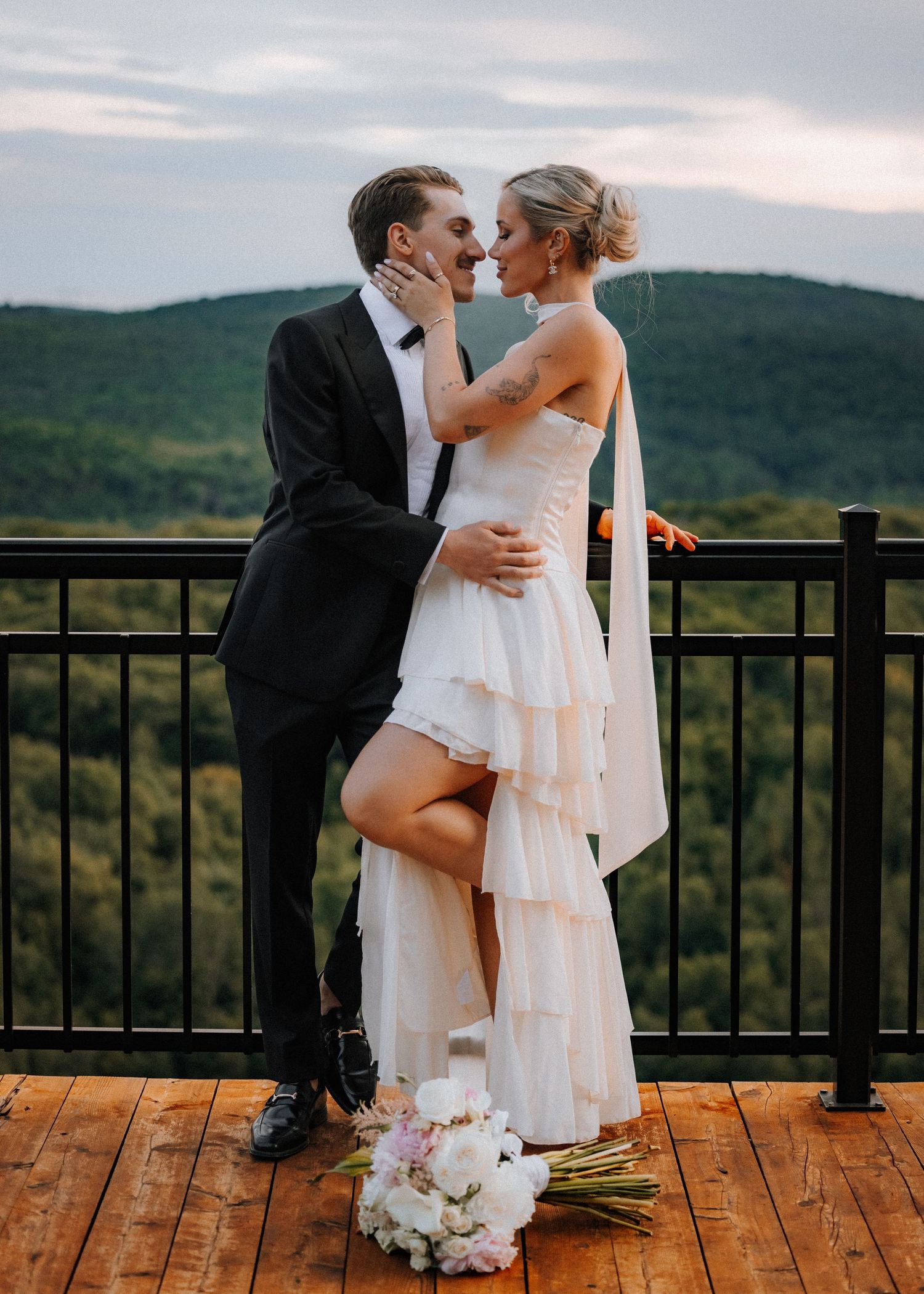 Bride and groom share a romantic kiss on a wooden deck overlooking lush green mountains, with a white bouquet at their feet.