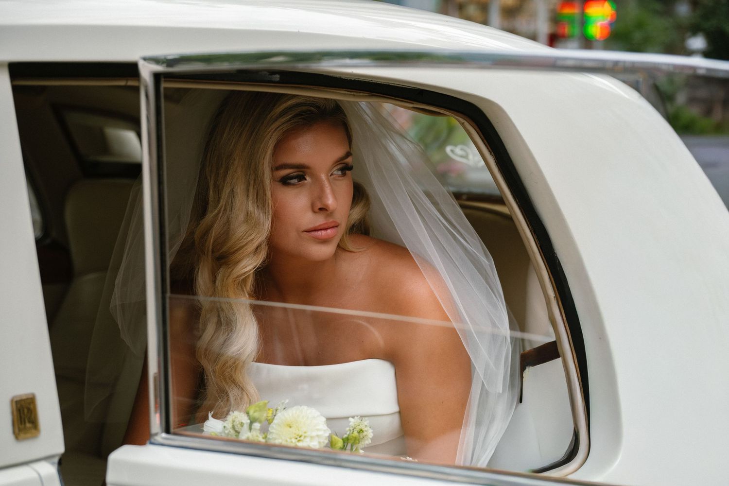 Blonde bride in white gown and veil sitting inside a white luxury limousine on her wedding day.