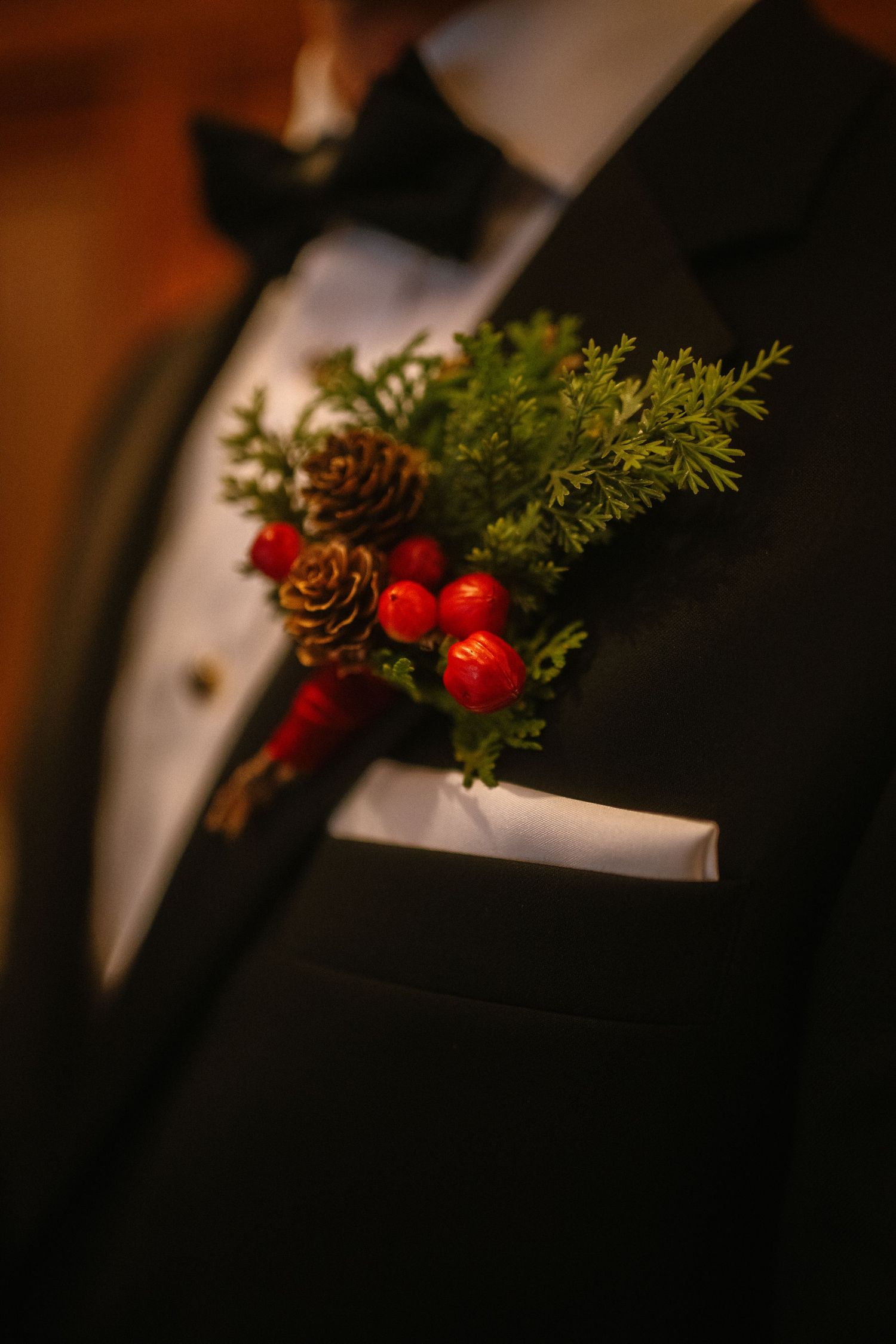 Close-up of a festive wedding boutonniere with pine cones, red berries, and greenery on a black tuxedo lapel.
