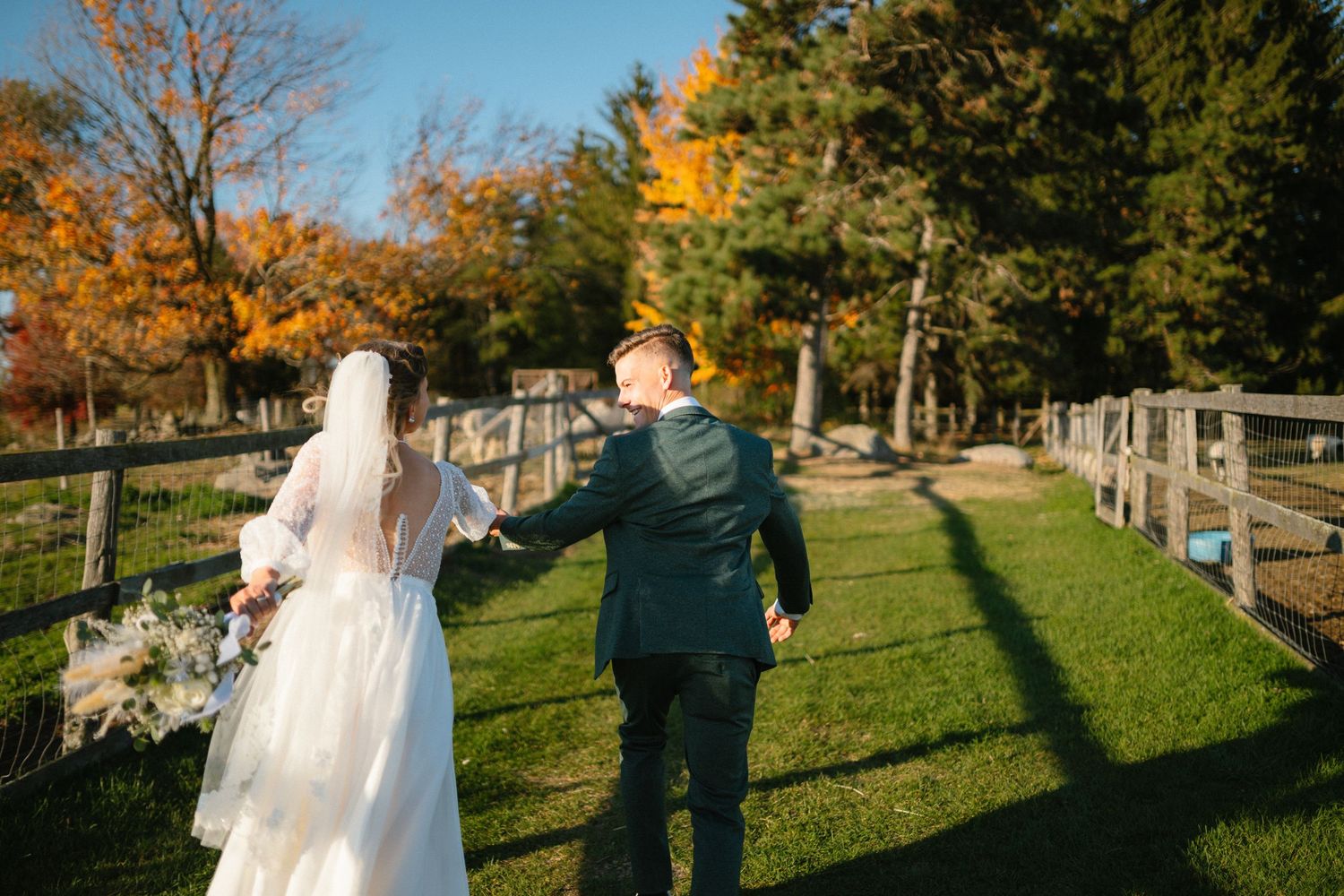 Bride and groom walking hand in hand along a wooden fence on a sunny autumn day at an outdoor wedding venue.