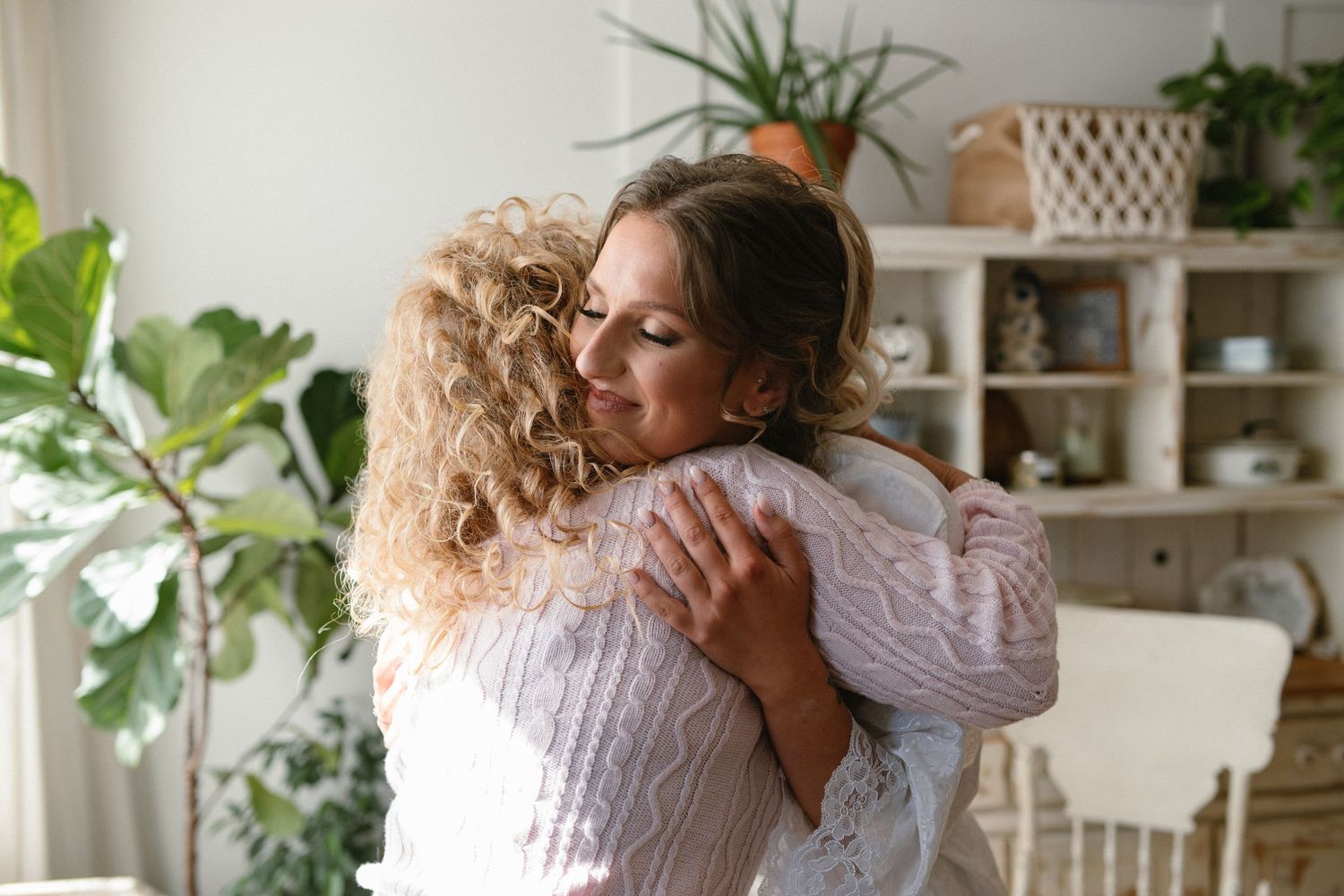 Two women sharing a warm embrace in a cozy, plant-filled living room with white decor and natural lighting.