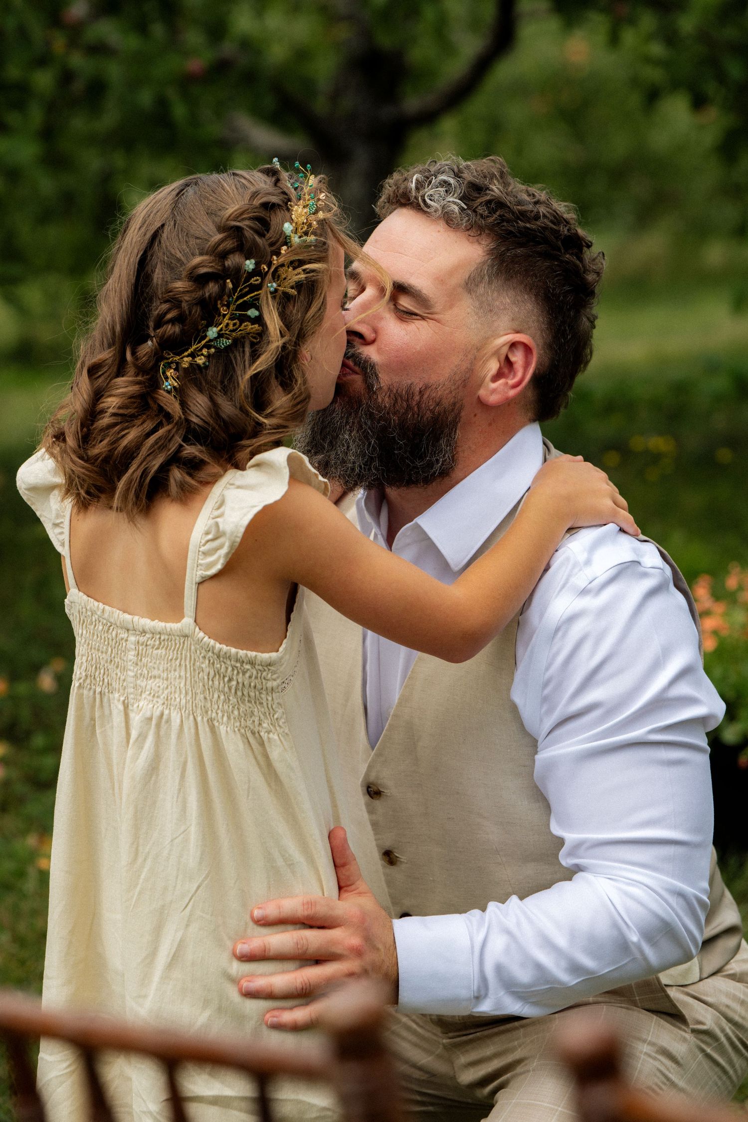 Bearded groom kisses young flower girl in cream dress with floral hair accessories in lush garden setting.