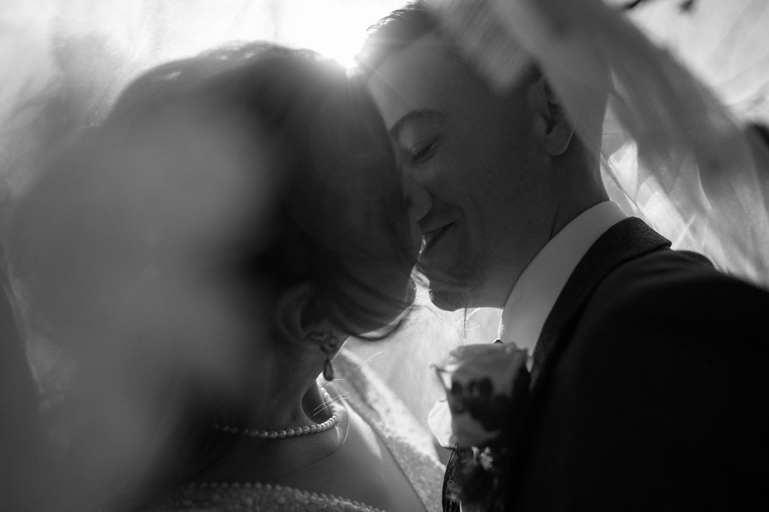 Black and white romantic close-up of bride and groom nearly kissing beneath wedding veil.