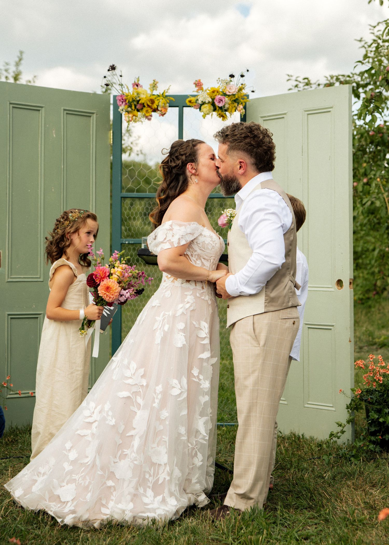 Bride and groom share a first kiss at outdoor wedding ceremony with green doors and floral arch backdrop.