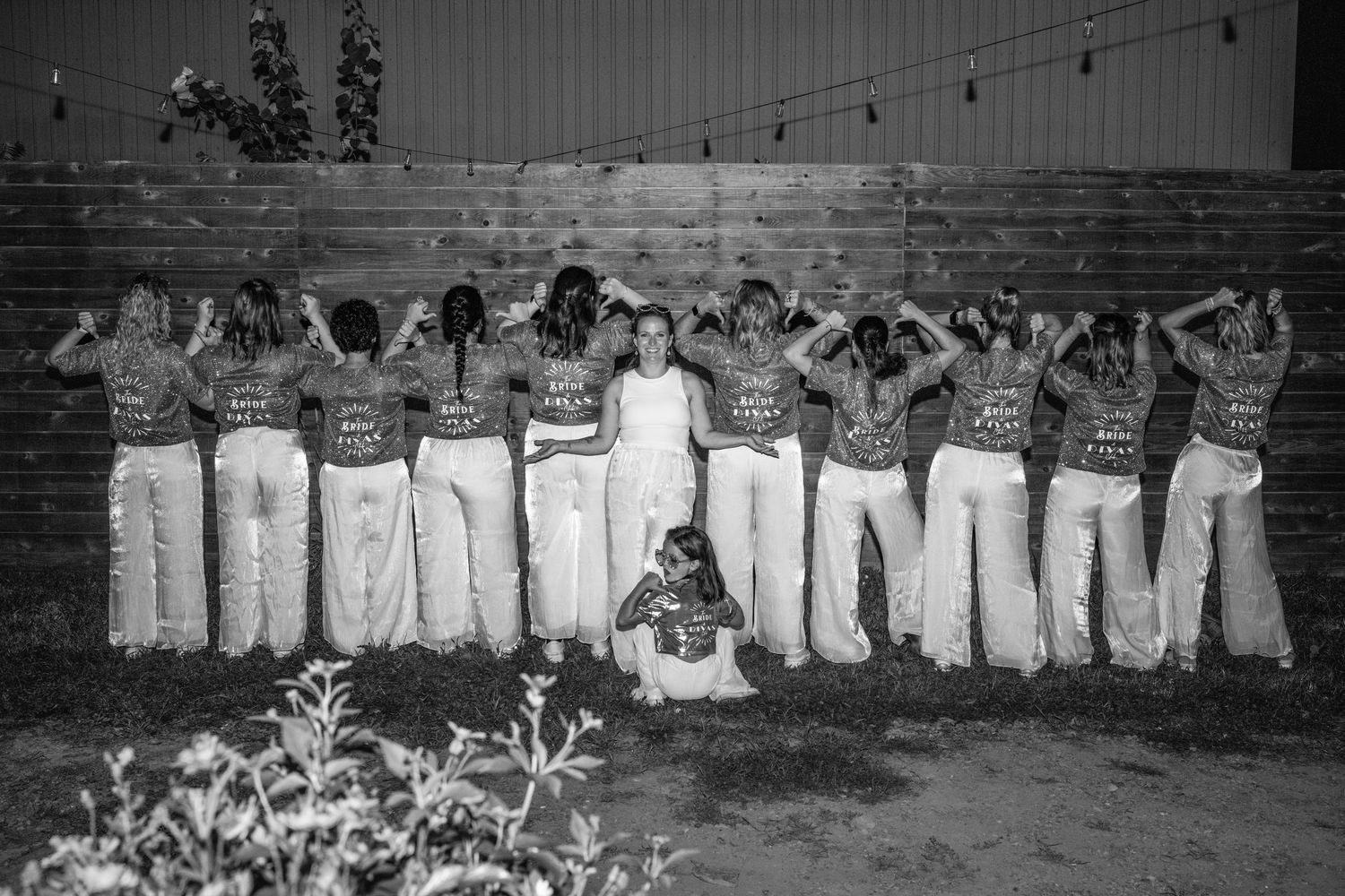 Bride poses with bridesmaids in matching tops and white pants, all facing away, in black and white outdoor photo.