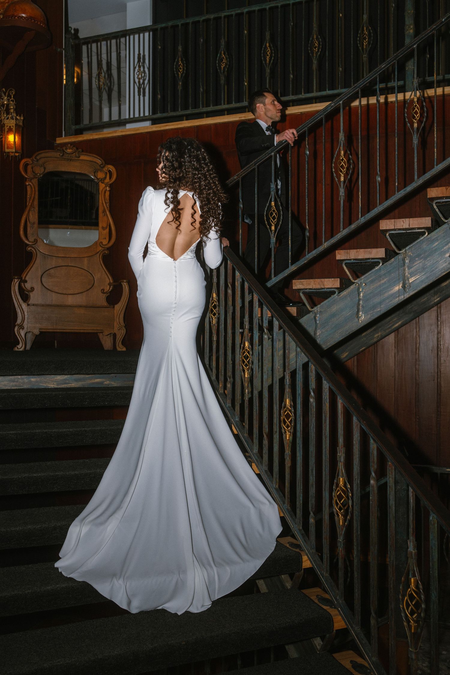 Bride in elegant backless white mermaid gown stands on grand staircase as groom watches from above.