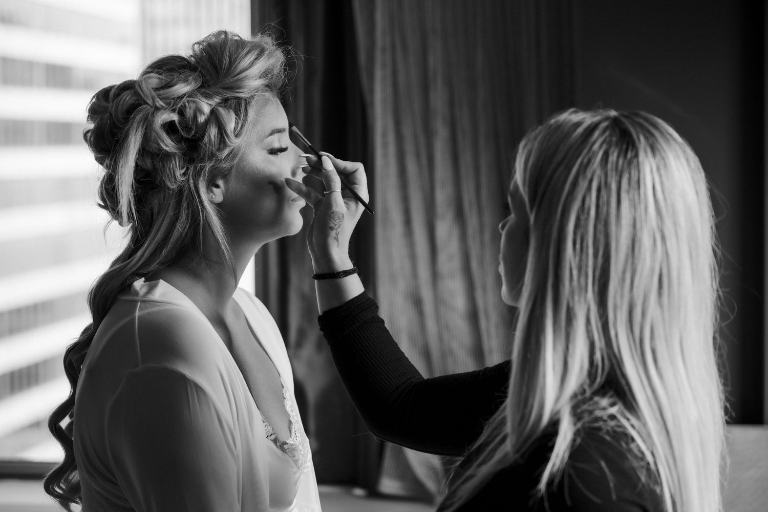 Black and white photo of a makeup artist applying makeup to a bride with elegant curled hair before her wedding.