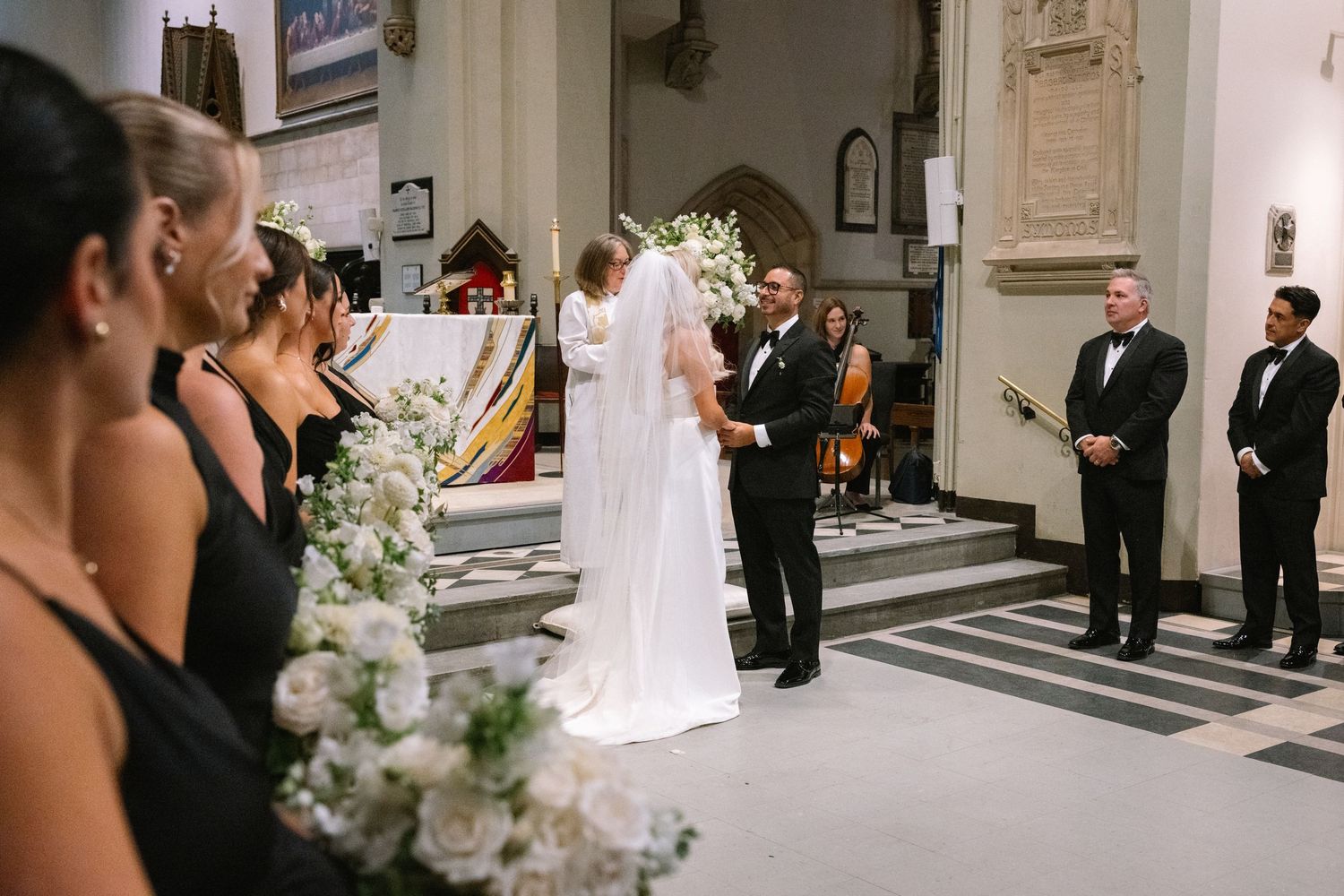 Bride and groom exchanging vows at church altar surrounded by bridesmaids and white floral arrangements.