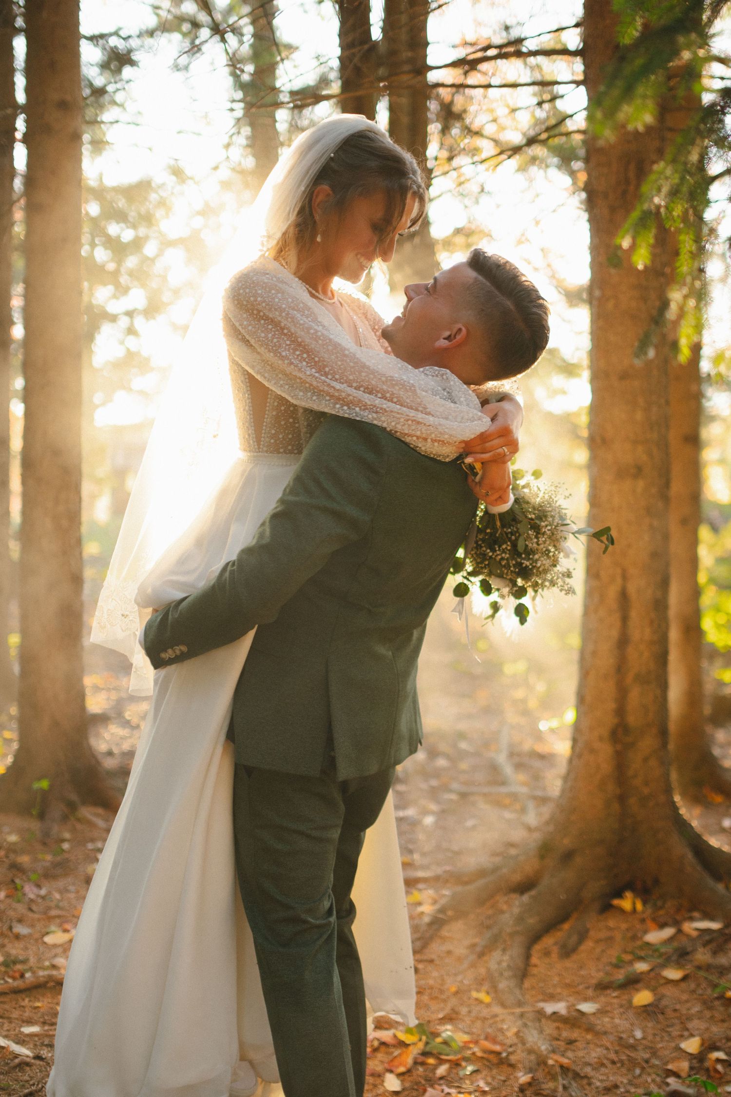 Groom lifting bride in sunlit forest during golden hour wedding portrait session.