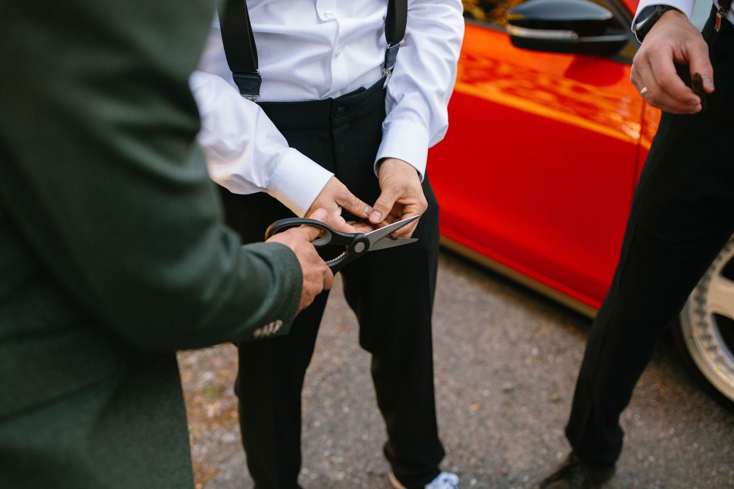 Groomsmen fastening cufflinks on a white dress shirt with black suspenders near a red car.