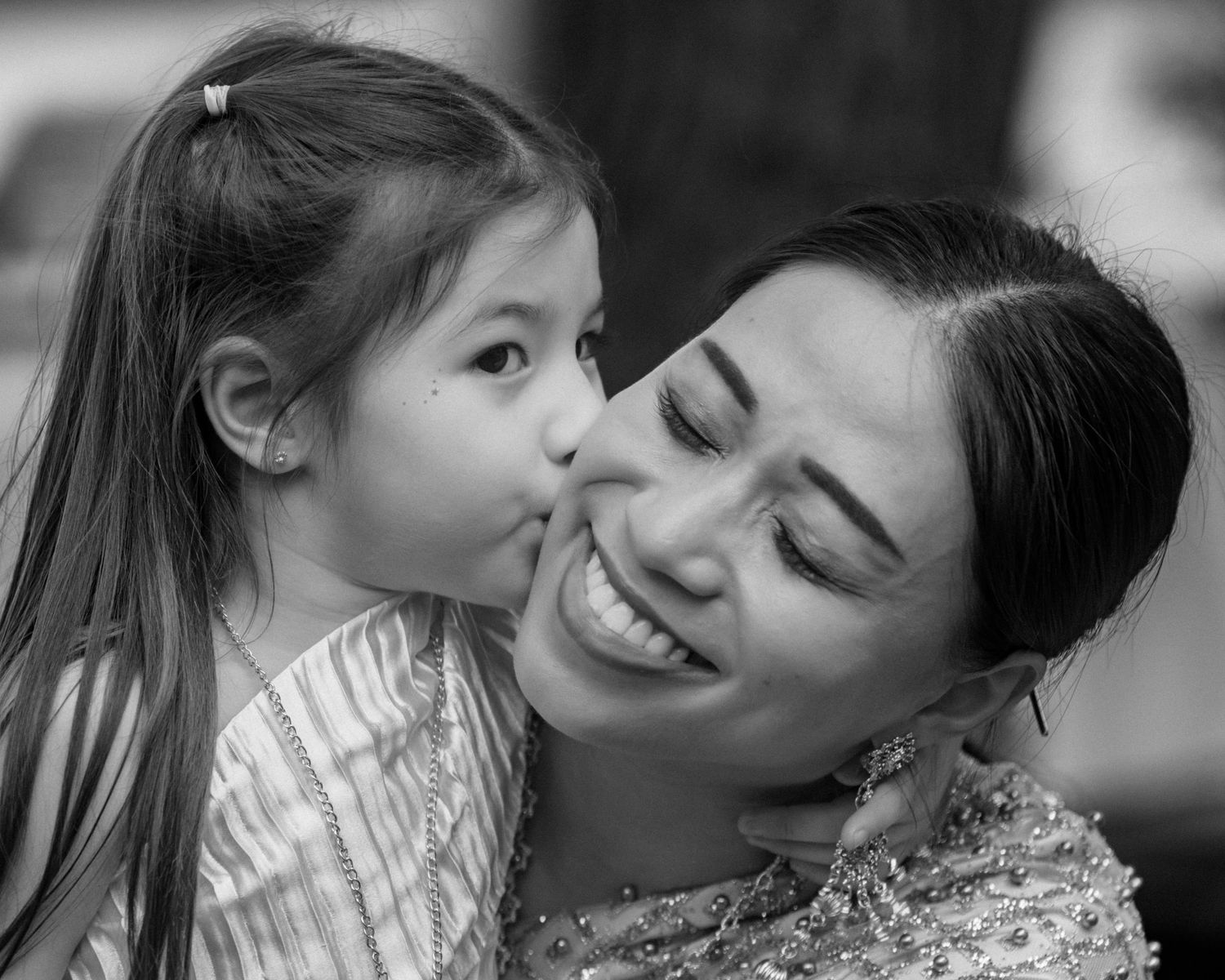 A young girl kisses her smiling mother on the cheek in a tender black and white portrait photo.