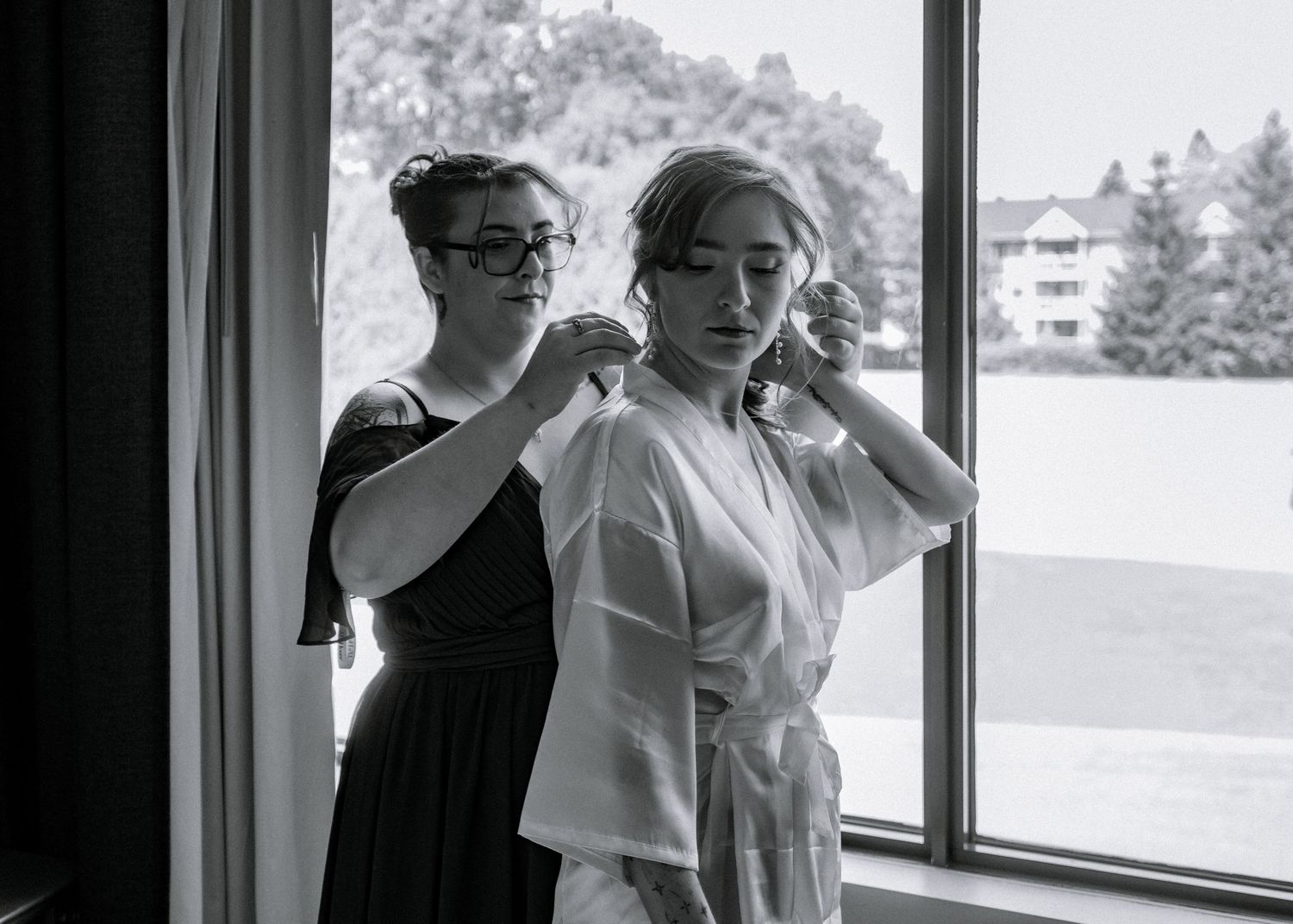 Bridesmaid helping bride with dress in black and white wedding preparation photo near window.