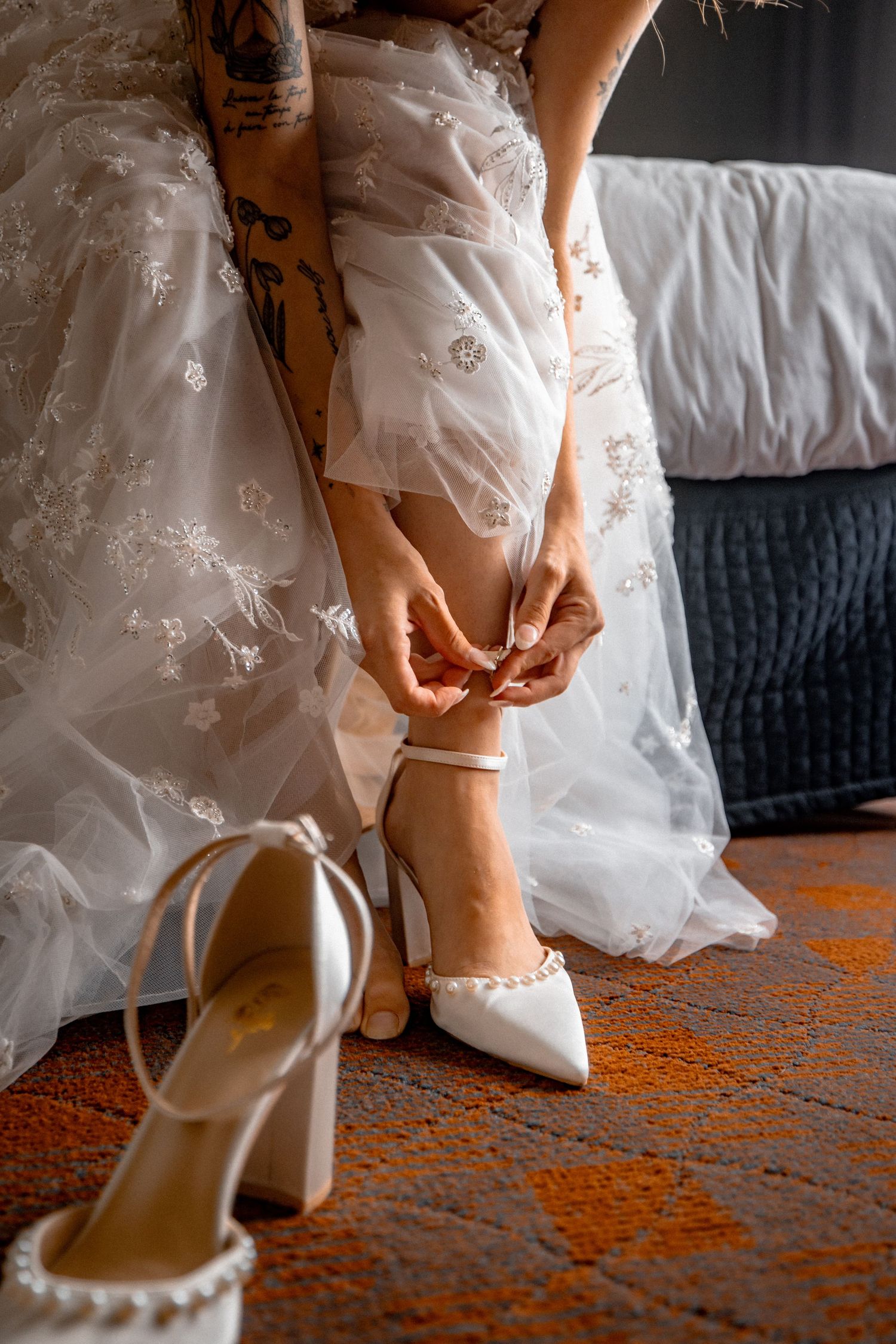 Bride fastening white block heel wedding shoes with pearl details while wearing a lace wedding gown.