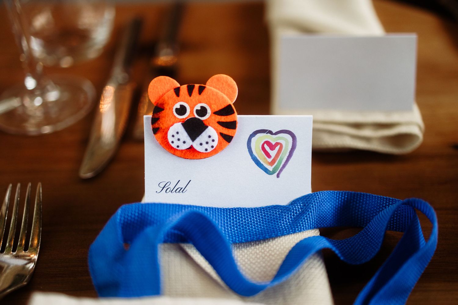 Tiger-shaped place card holder with name Solal on a table setting with blue ribbon and heart drawing.