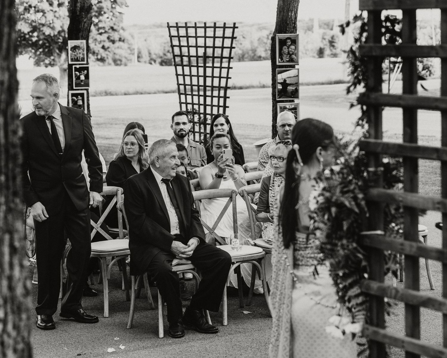Black and white photo of wedding guests seated outdoors during a ceremony, with a decorative trellis backdrop.
