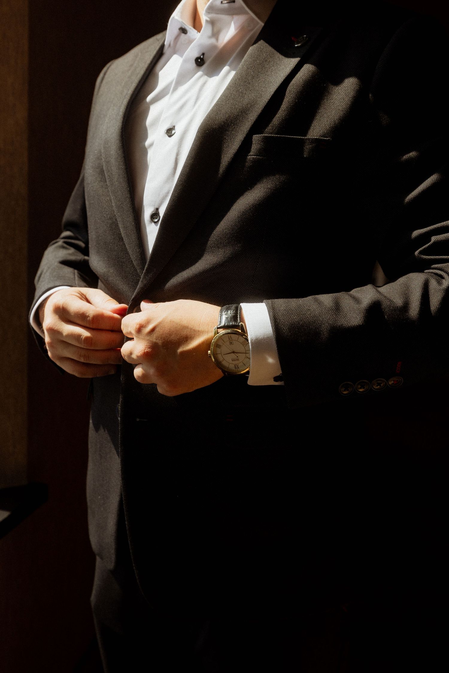 Groom in black suit buttoning jacket with classic watch on wrist in dramatic lighting.