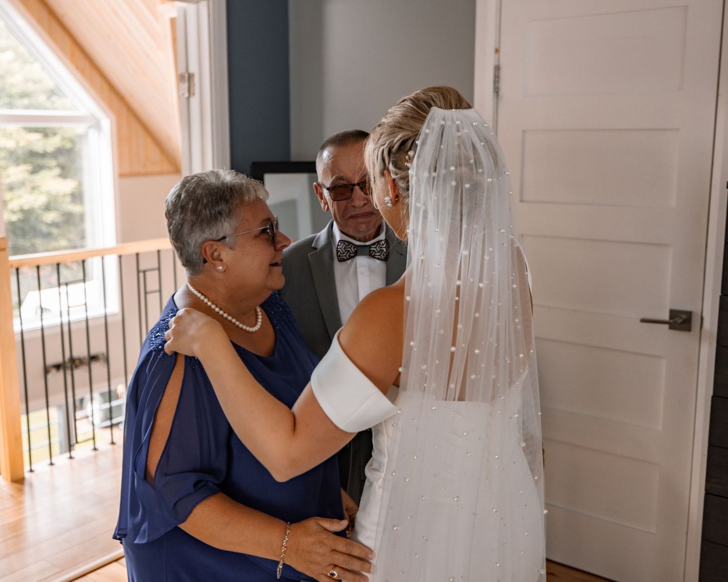Bride in white gown and veil shares an emotional moment with her parents in a blue dress and suit before the wedding ceremony.