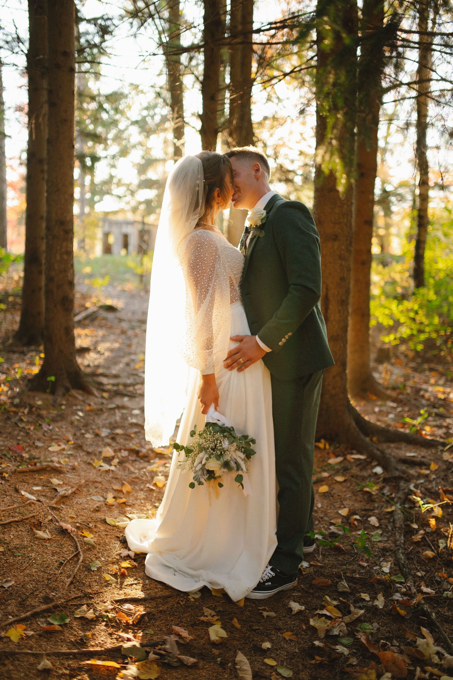 Bride and groom kissing among tall trees in a sun-drenched autumn forest setting.