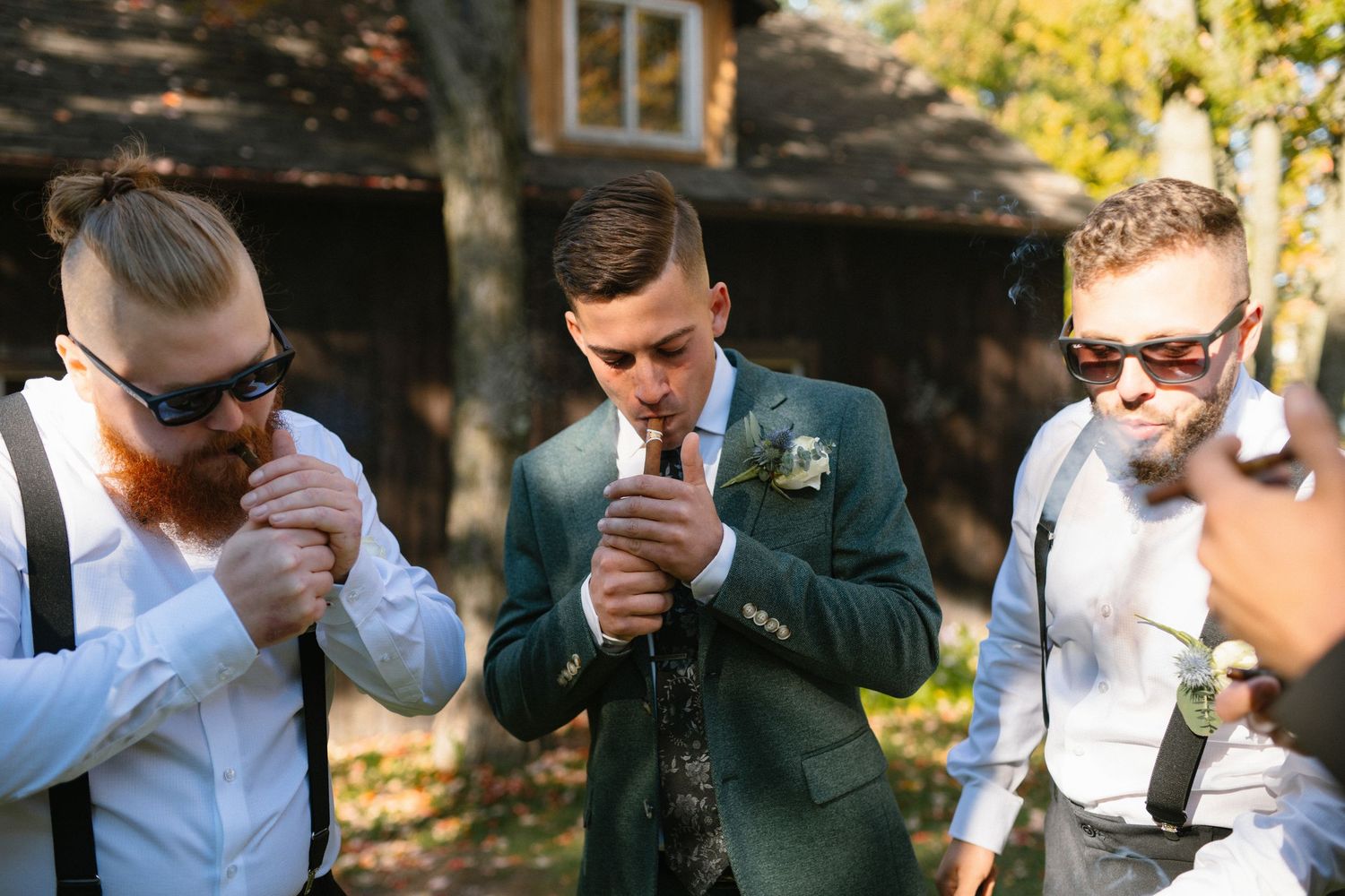 Groomsmen in sunglasses lighting cigars outdoors near a rustic cabin on a wedding day.