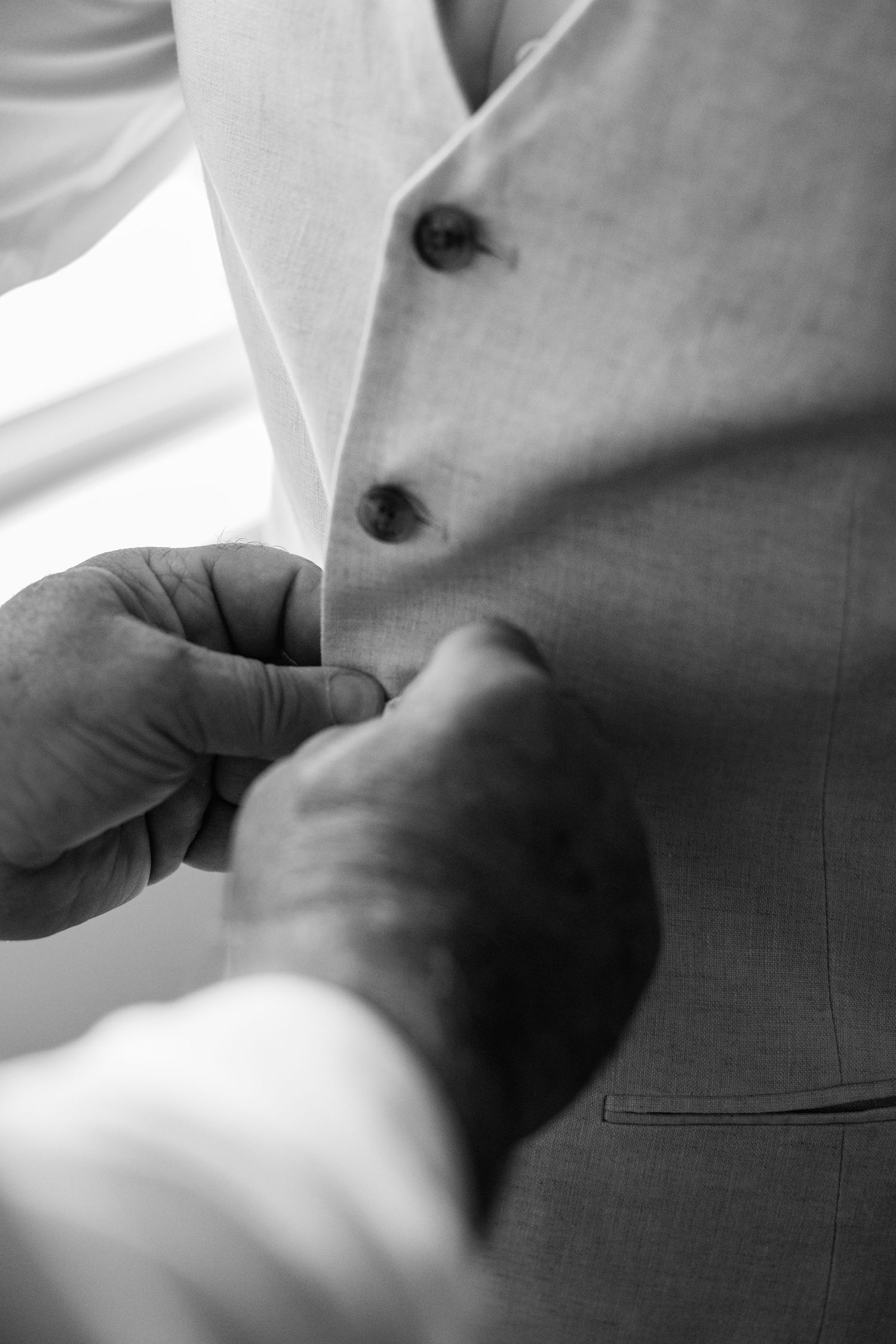 Close-up black and white photo of hands buttoning a groom's wedding vest.